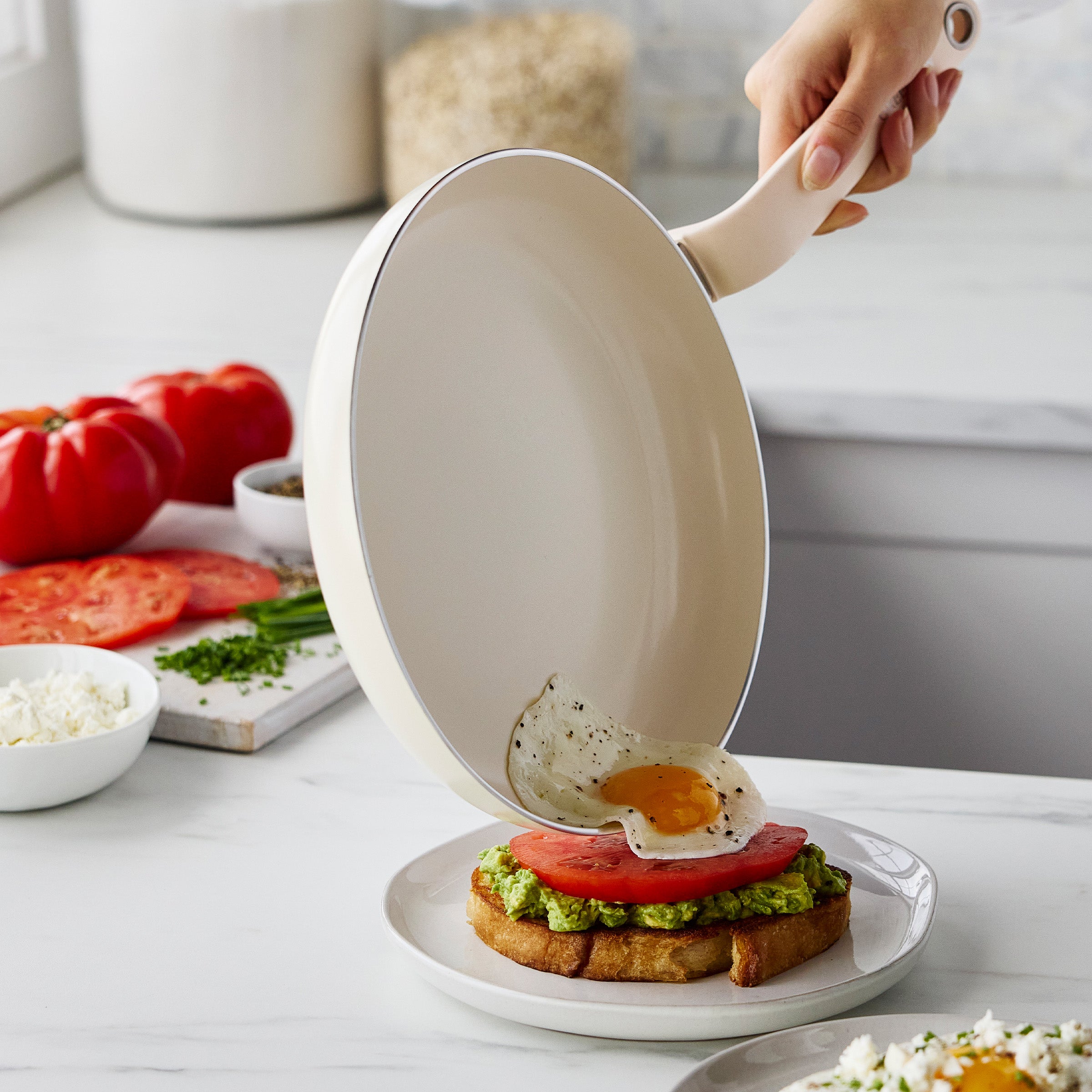 A hand slides a fried egg from a cream-colored ceramic nonstick frypan onto avocado toast with tomato on a plate.