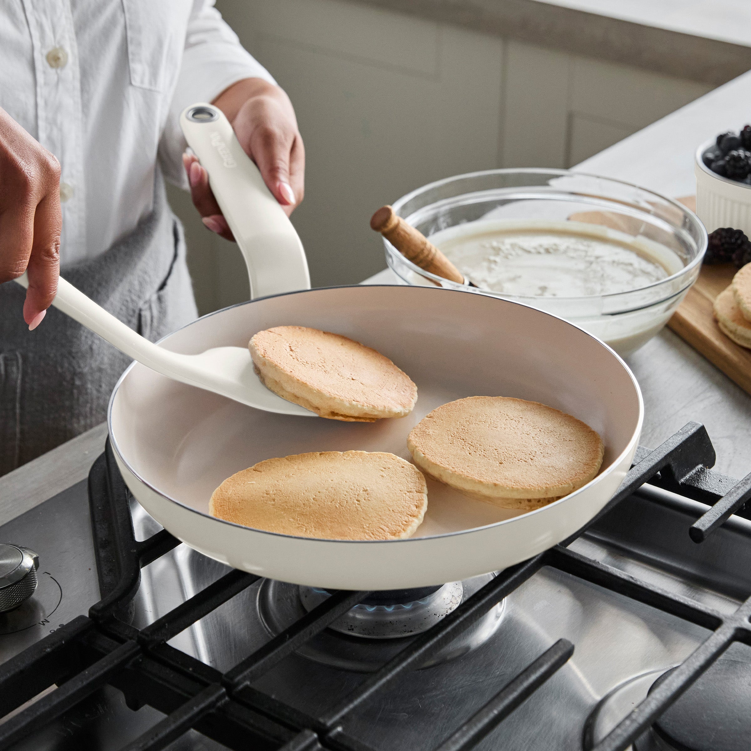 Pancakes flipping in a cream ceramic nonstick frypan, with bowls of batter and berries nearby on the counter.