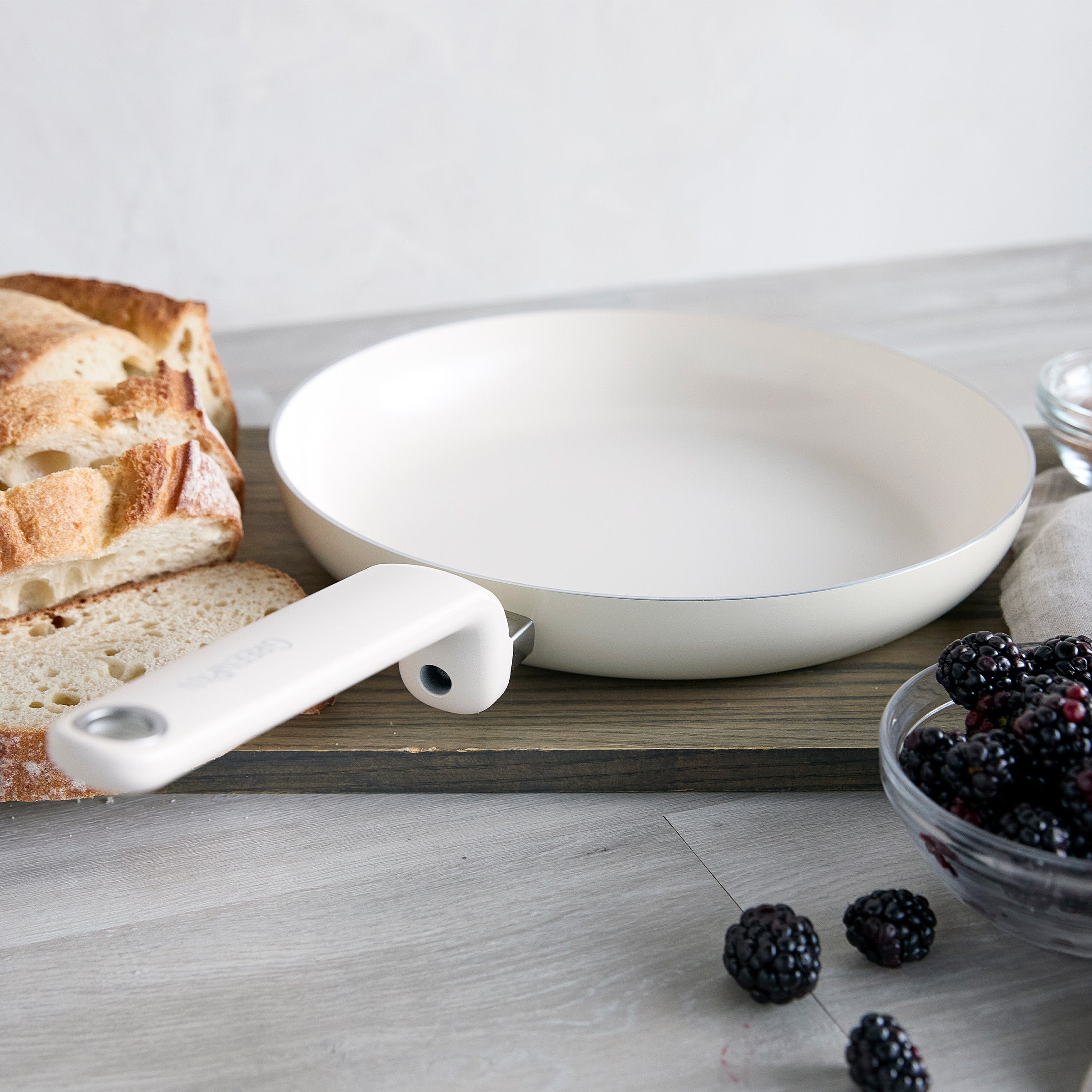 Cream ceramic nonstick frypan on a board next to sliced bread and a bowl of blackberries.