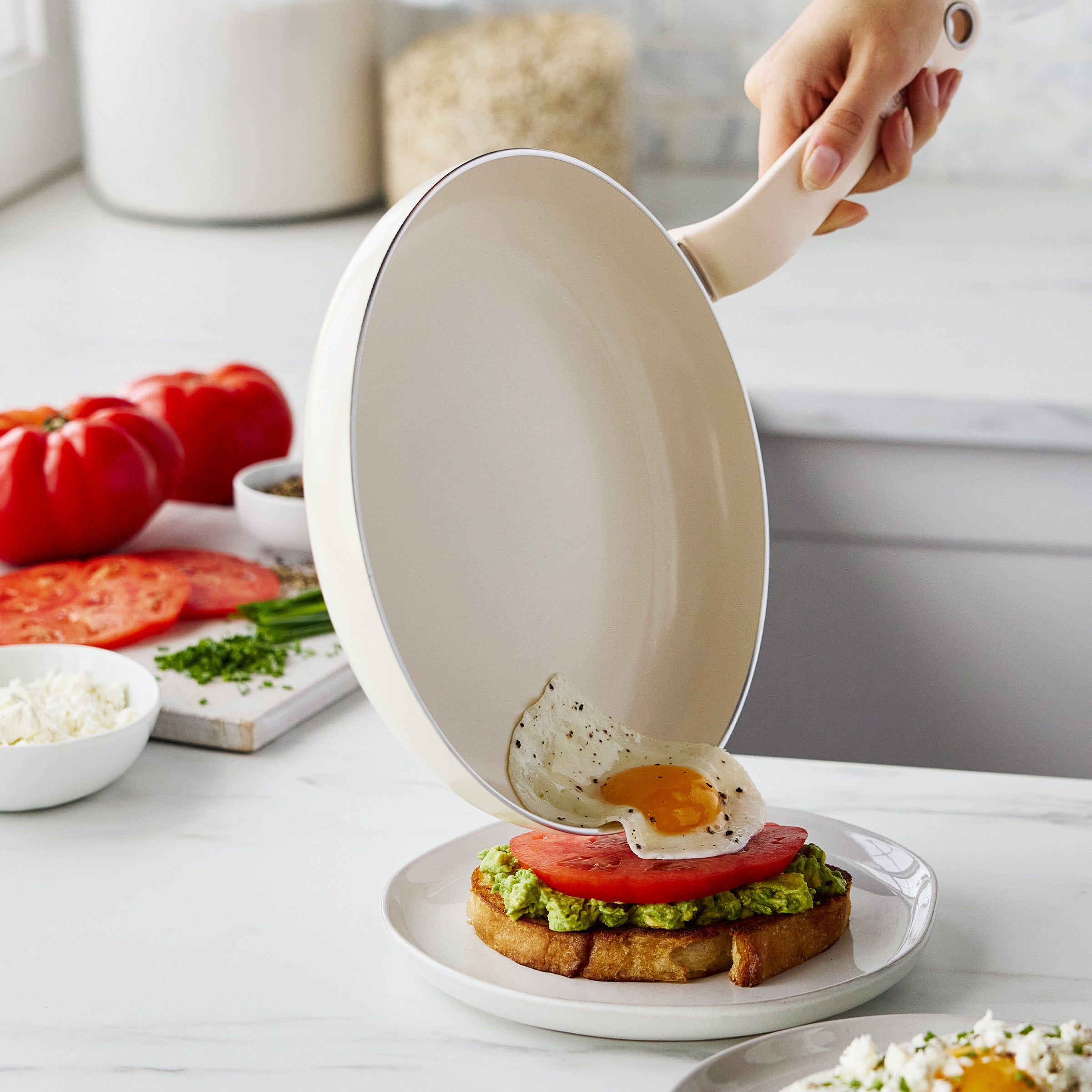 A hand slides a fried egg from a cream ceramic frypan onto avocado toast in a bright kitchen.