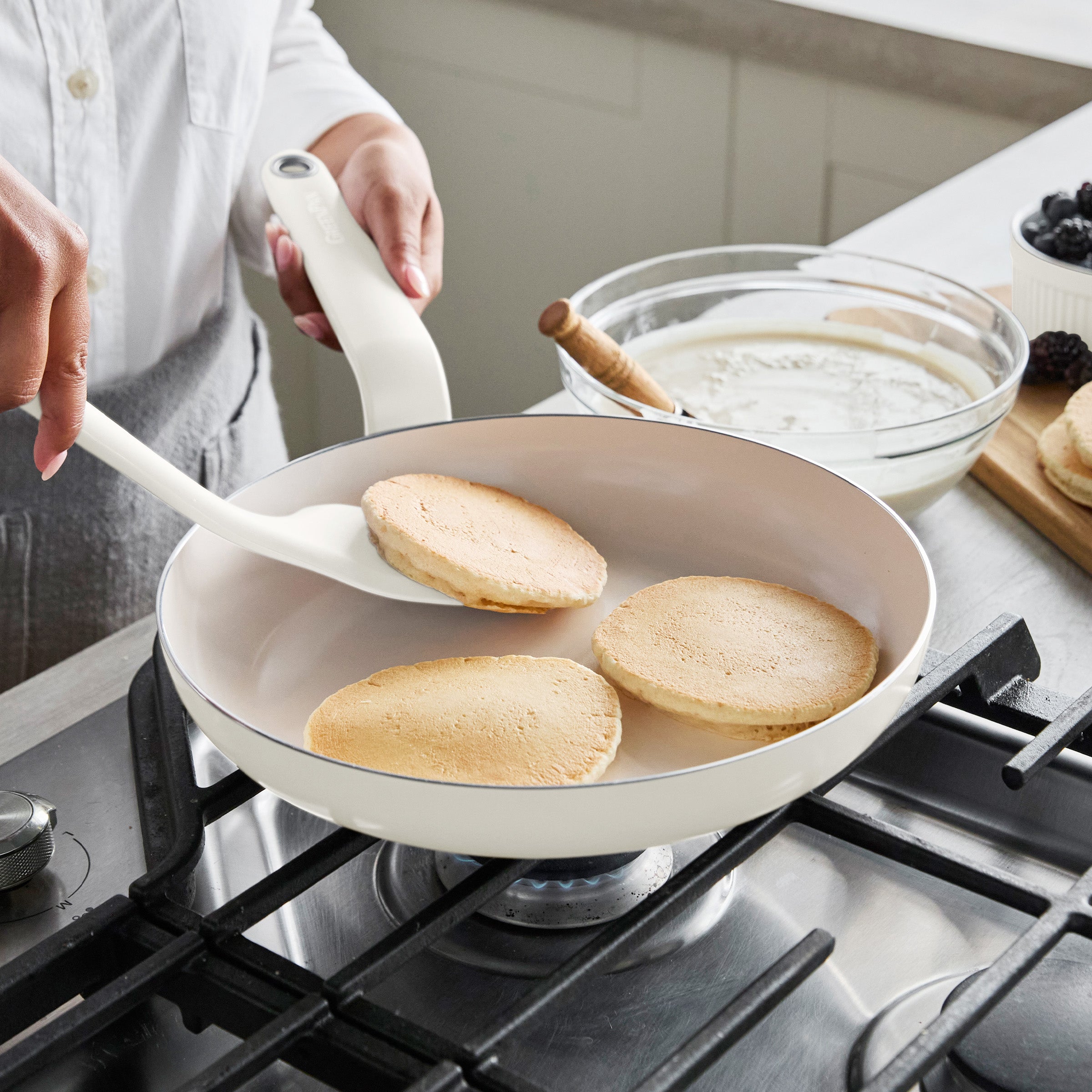 Flipping pancakes in a cream Rio Advanced Ceramic Nonstick 10 frypan, with pancake batter and berries on the counter nearby.