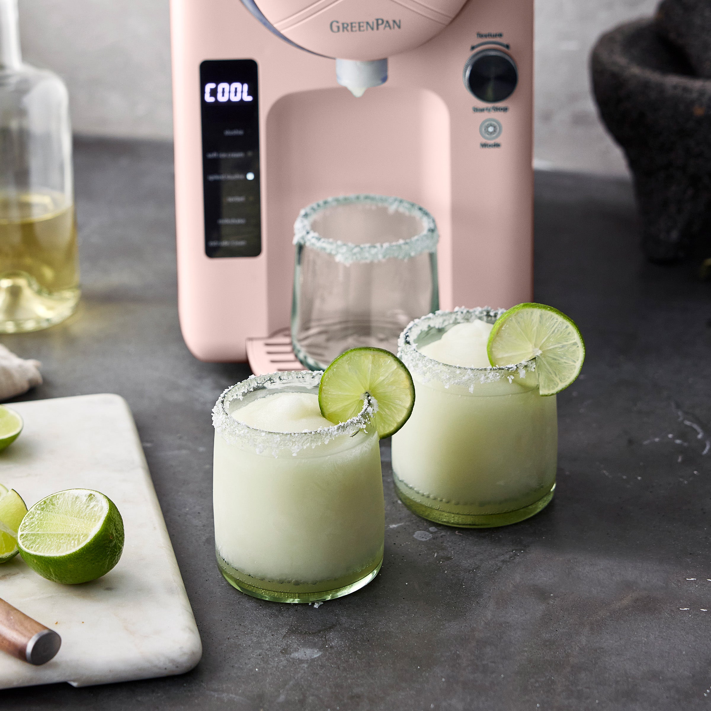 Two margaritas with lime wedges on a countertop next to a pink frozen treat maker