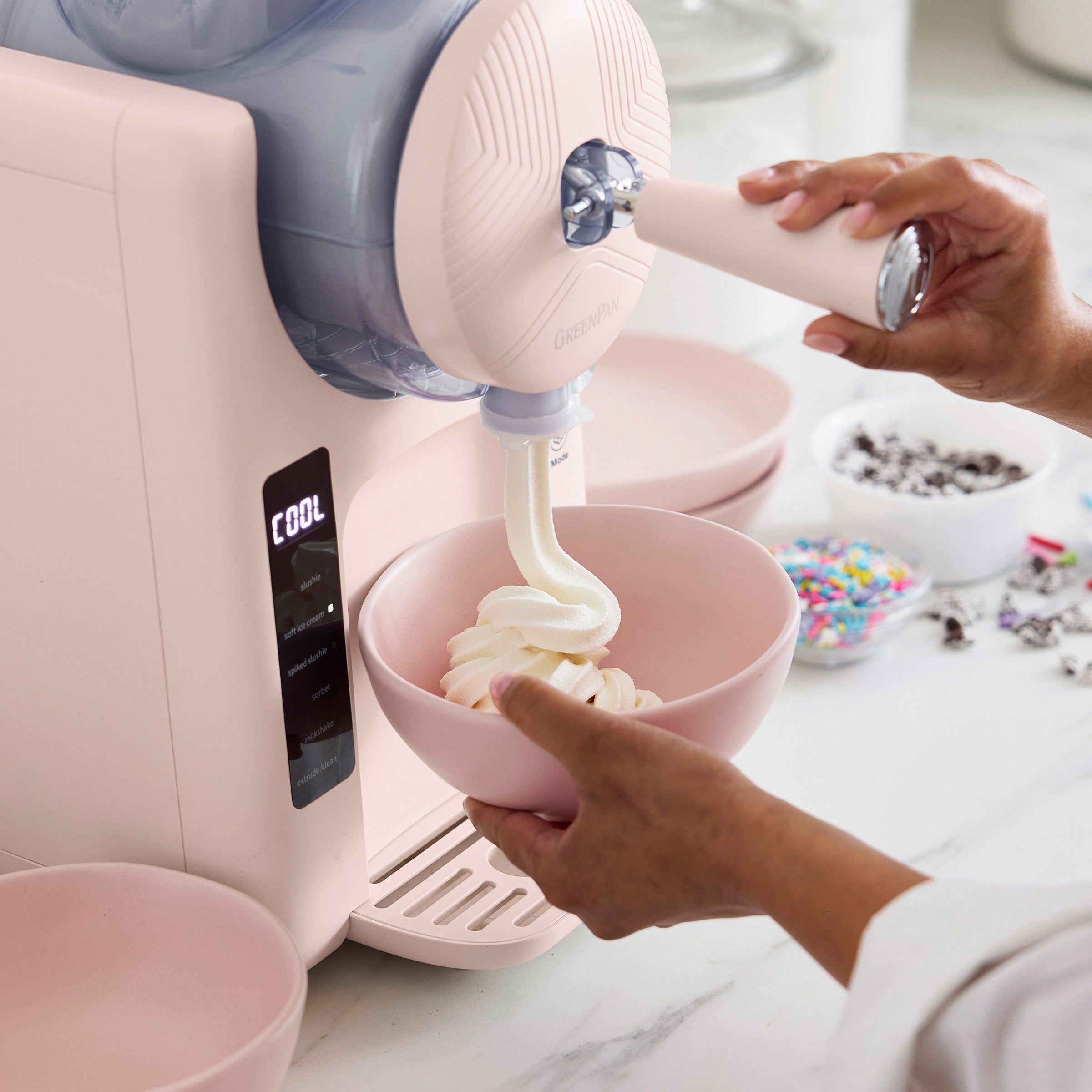Pink ice cream maker with a bowl of ice cream being filled, on a light-colored surface.