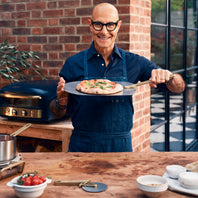 Man holding a pizza on a wooden table with a grill and ingredients in the background.