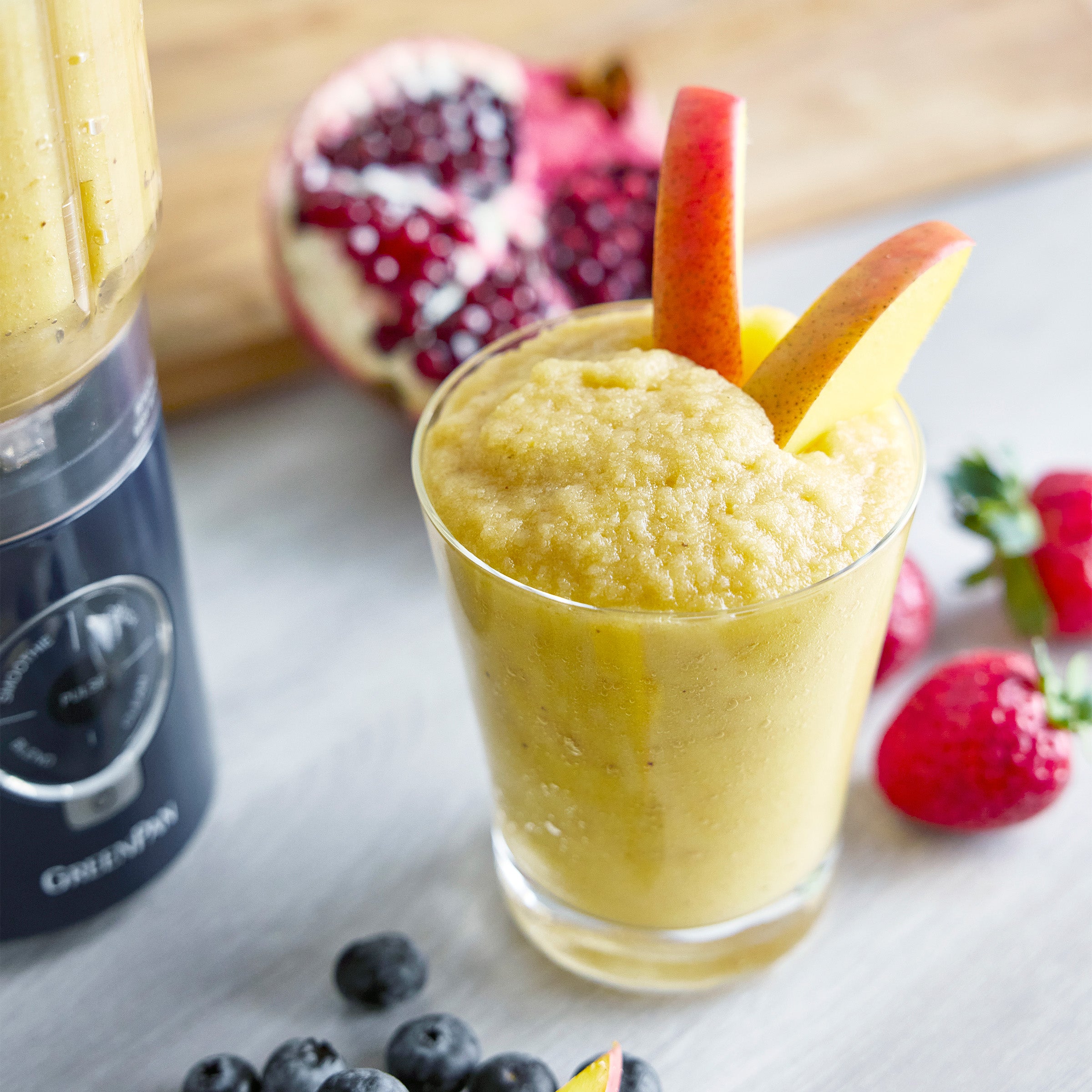 Yellow smoothie in a glass with fruit garnishes, next to a blender and fresh fruits on a light surface.