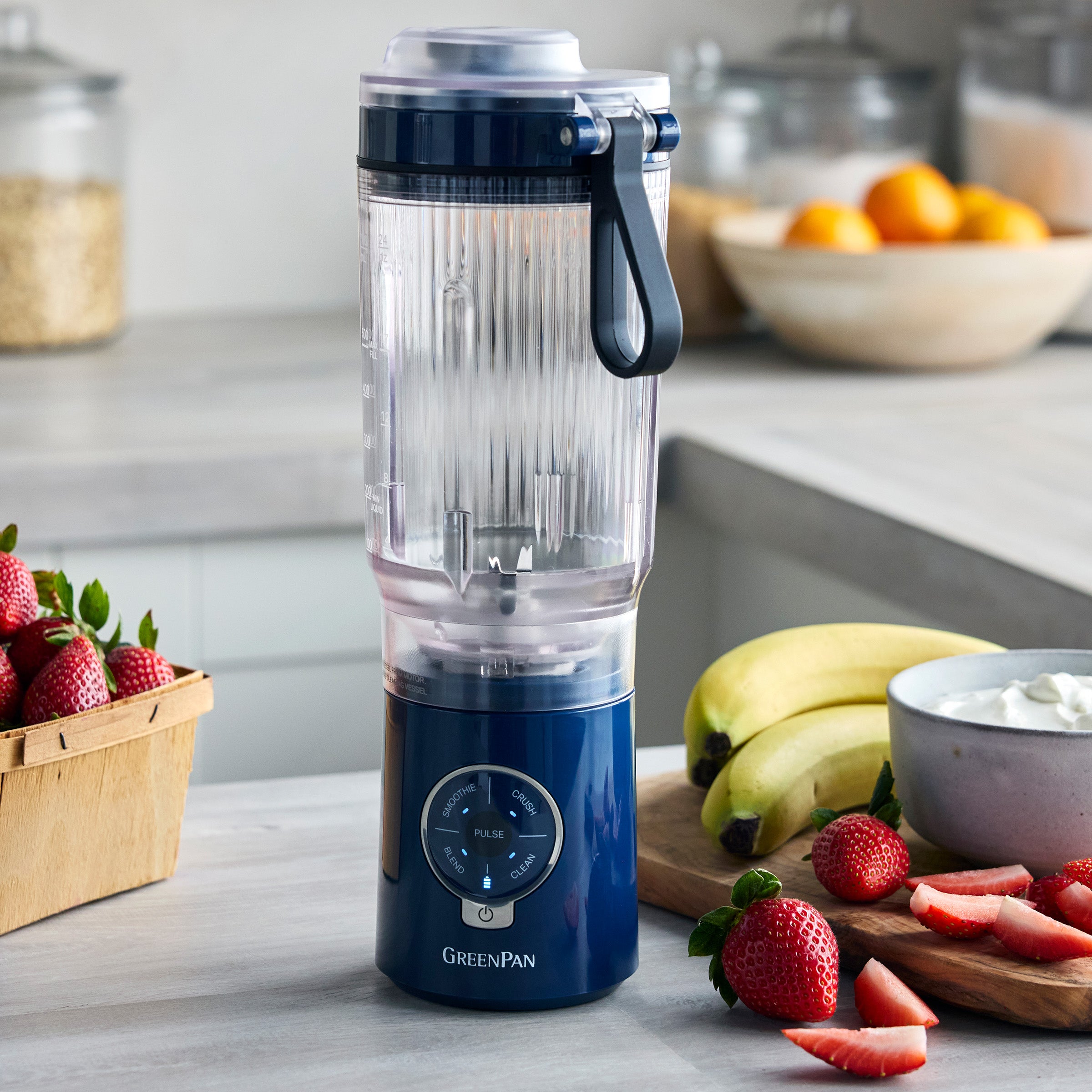 Blue portable blender on a kitchen counter with fruits and a bowl of yogurt.