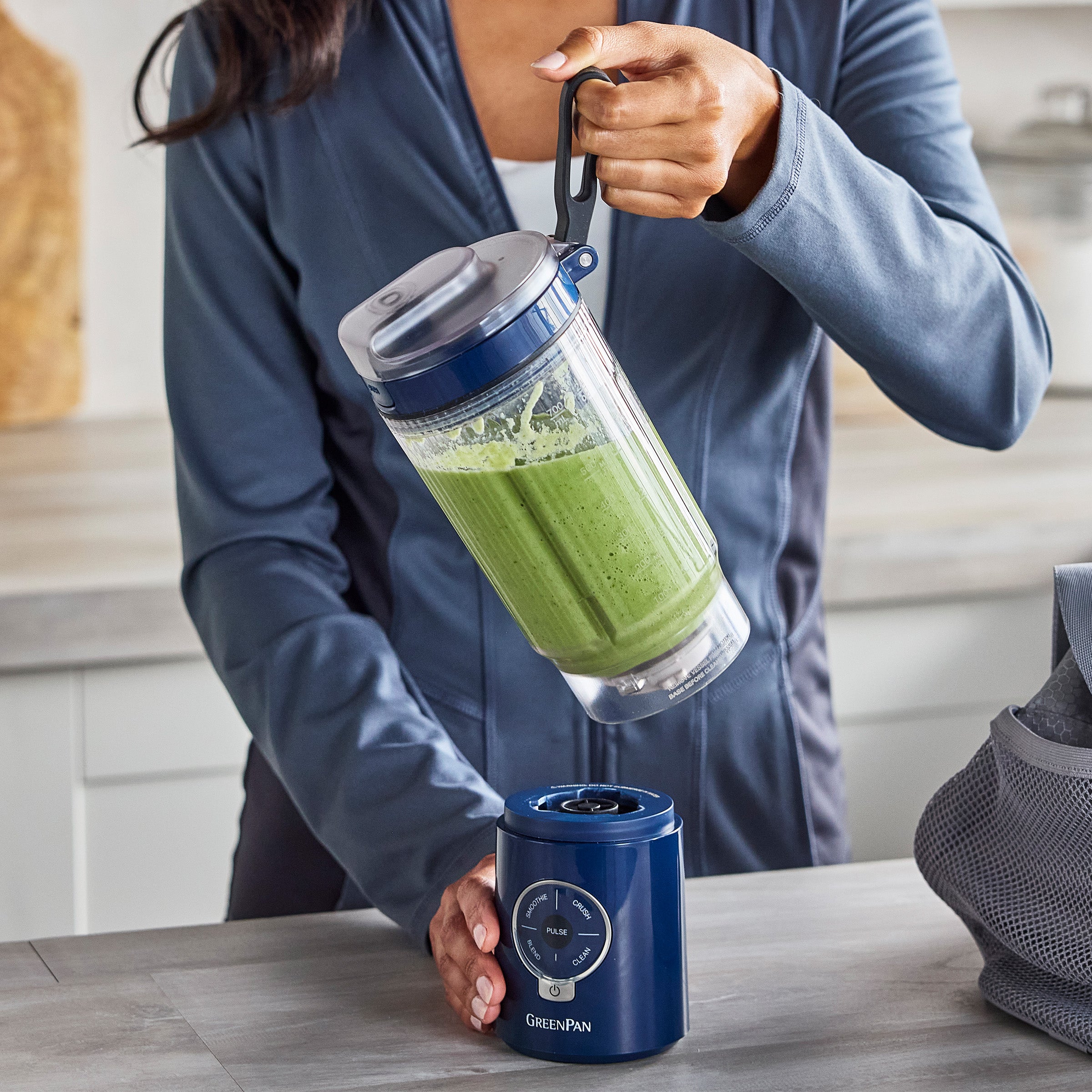 Person holding a GreenPan blender with a green smoothie inside, standing in a kitchen.