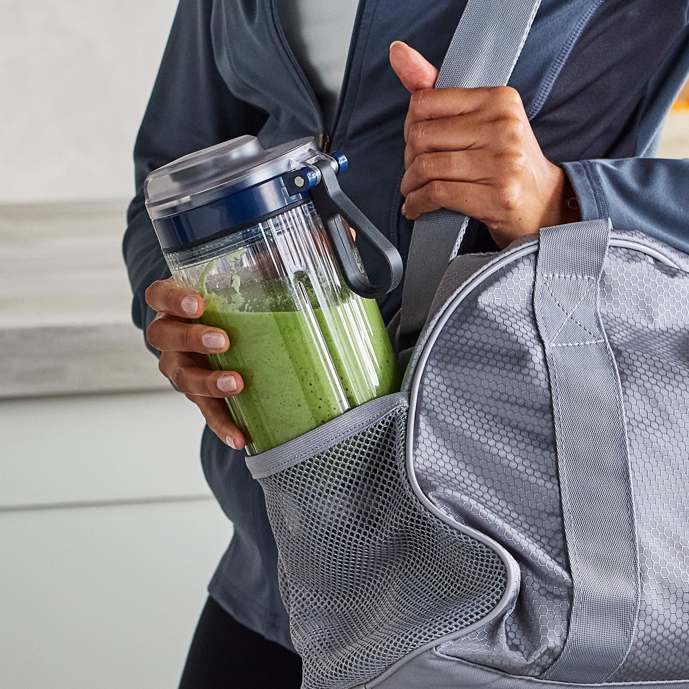 Person holding a green smoothie in a clear container with a blue lid, standing next to a gray bag.