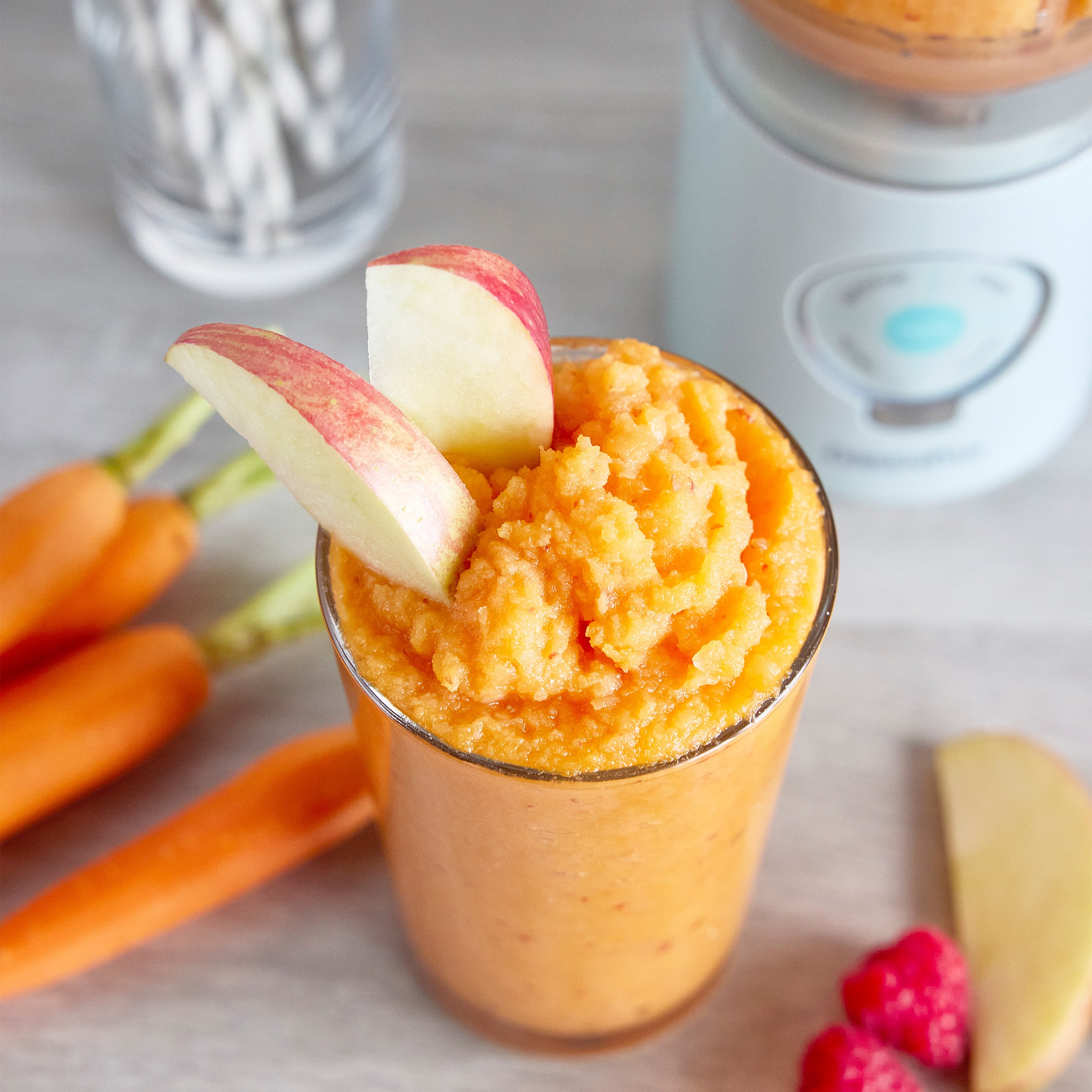 Orange smoothie in a glass with apple slices, carrots, and raspberries on a light surface