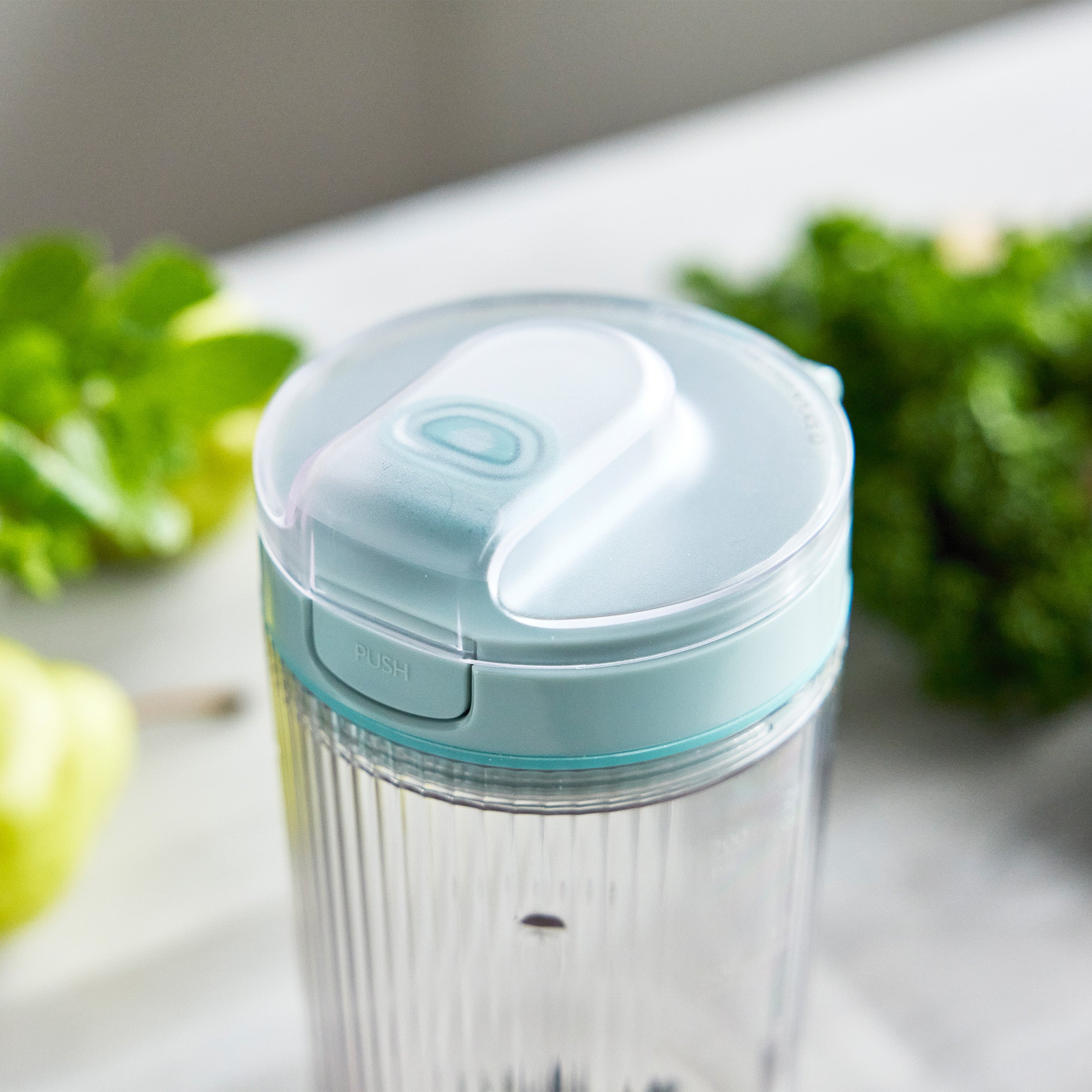 Clear blender container with blue lid on a light surface with green vegetables in the background