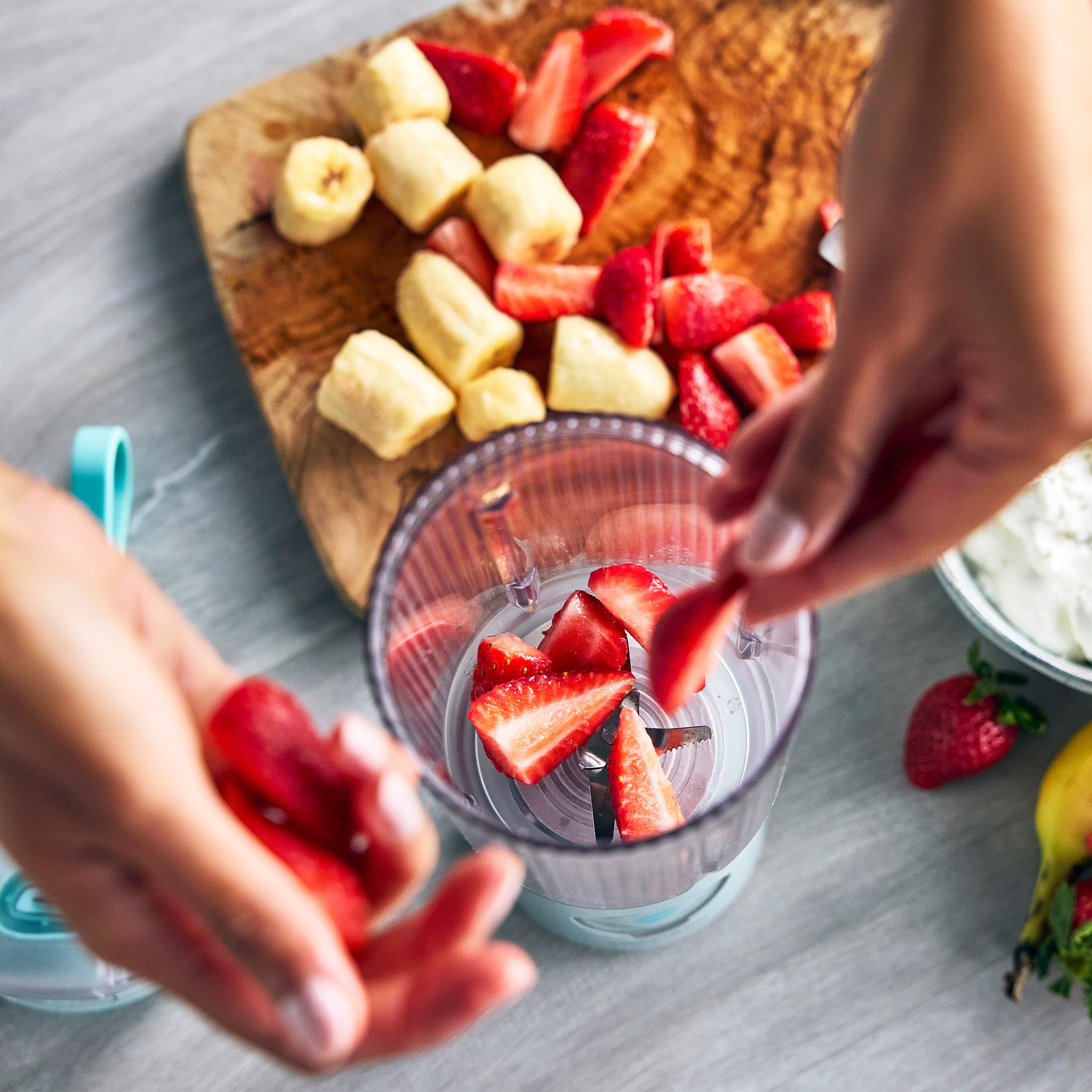 Person adding strawberries into a blender with a cutting board of sliced fruits in the background.