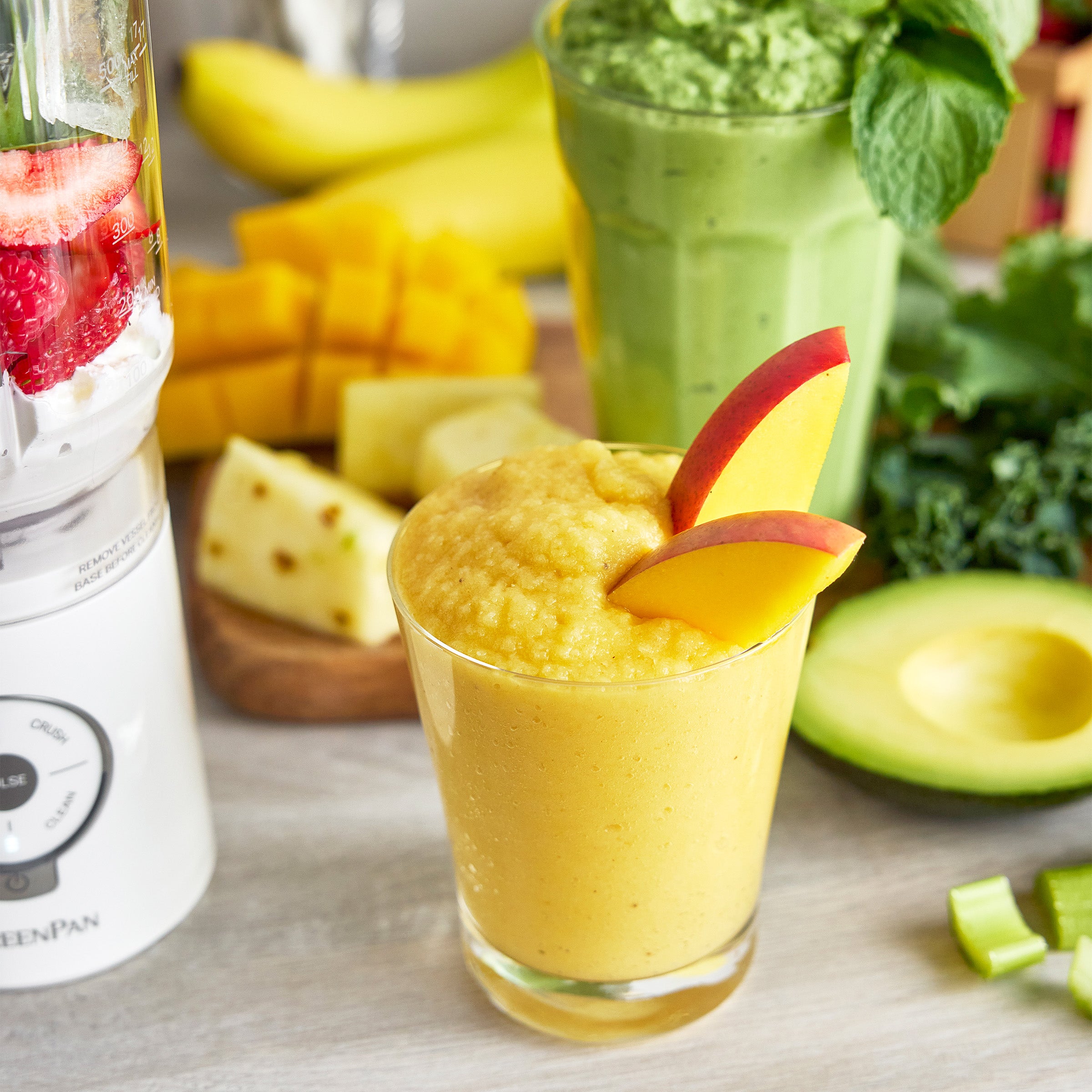 Yellow smoothie with a mango slice in a glass, blender, and fruits on a table.