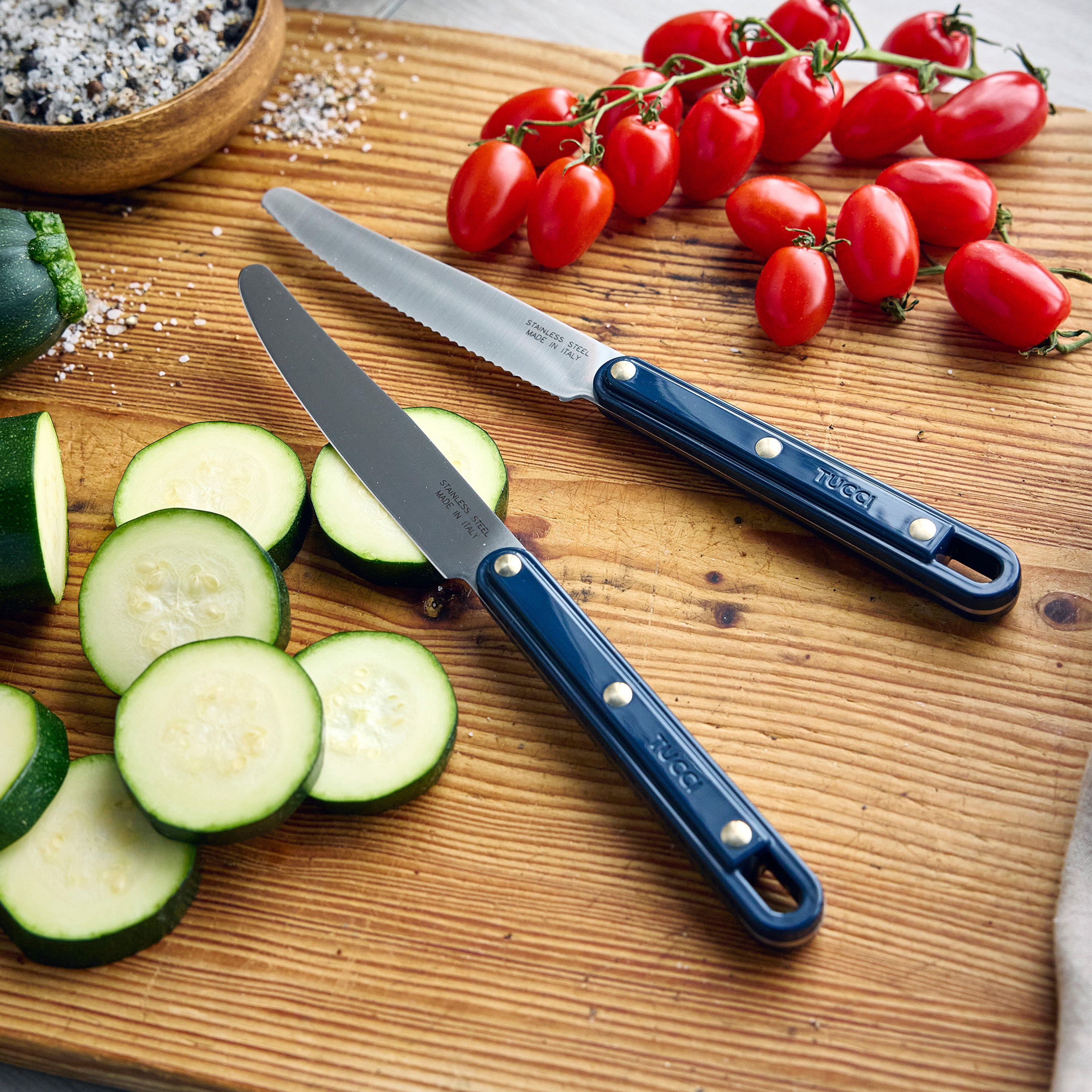 Two knives with blue handles on a wooden cutting board with sliced zucchini and tomatoes.