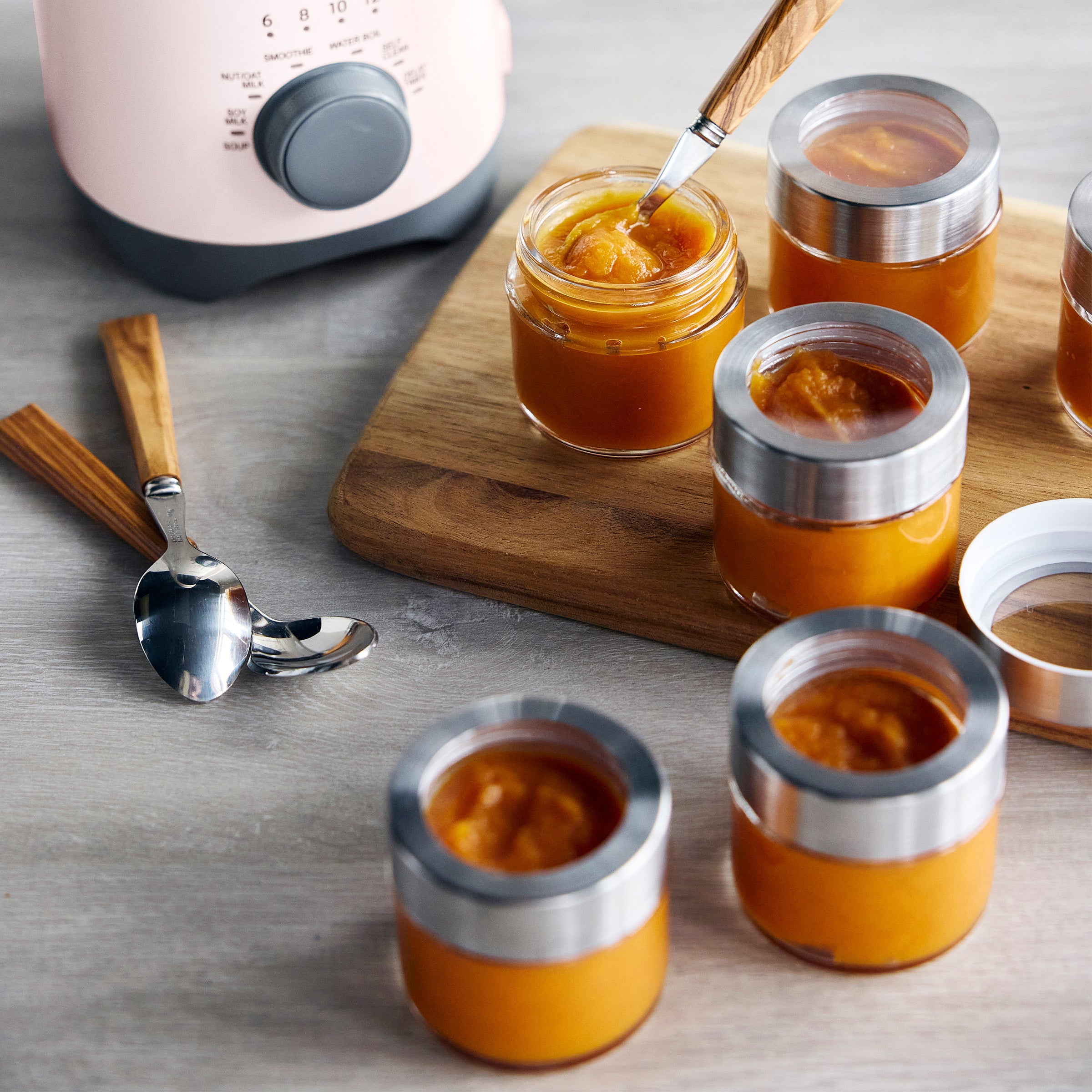 Small jars of baby food on a wooden board with spoons, next to a kitchen appliance.