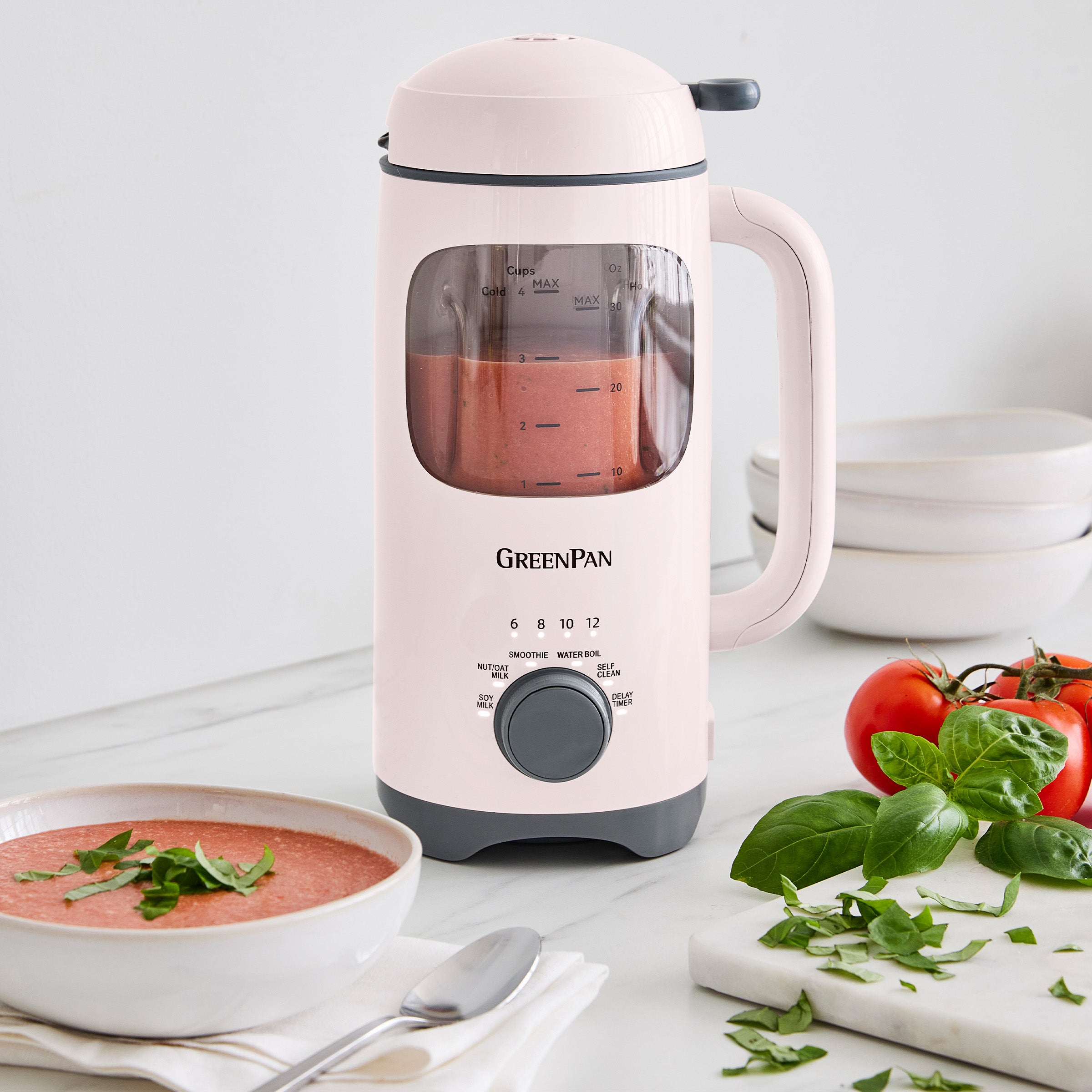 Pink GreenPan milk maker with a bowl of soup and ingredients on a white surface
