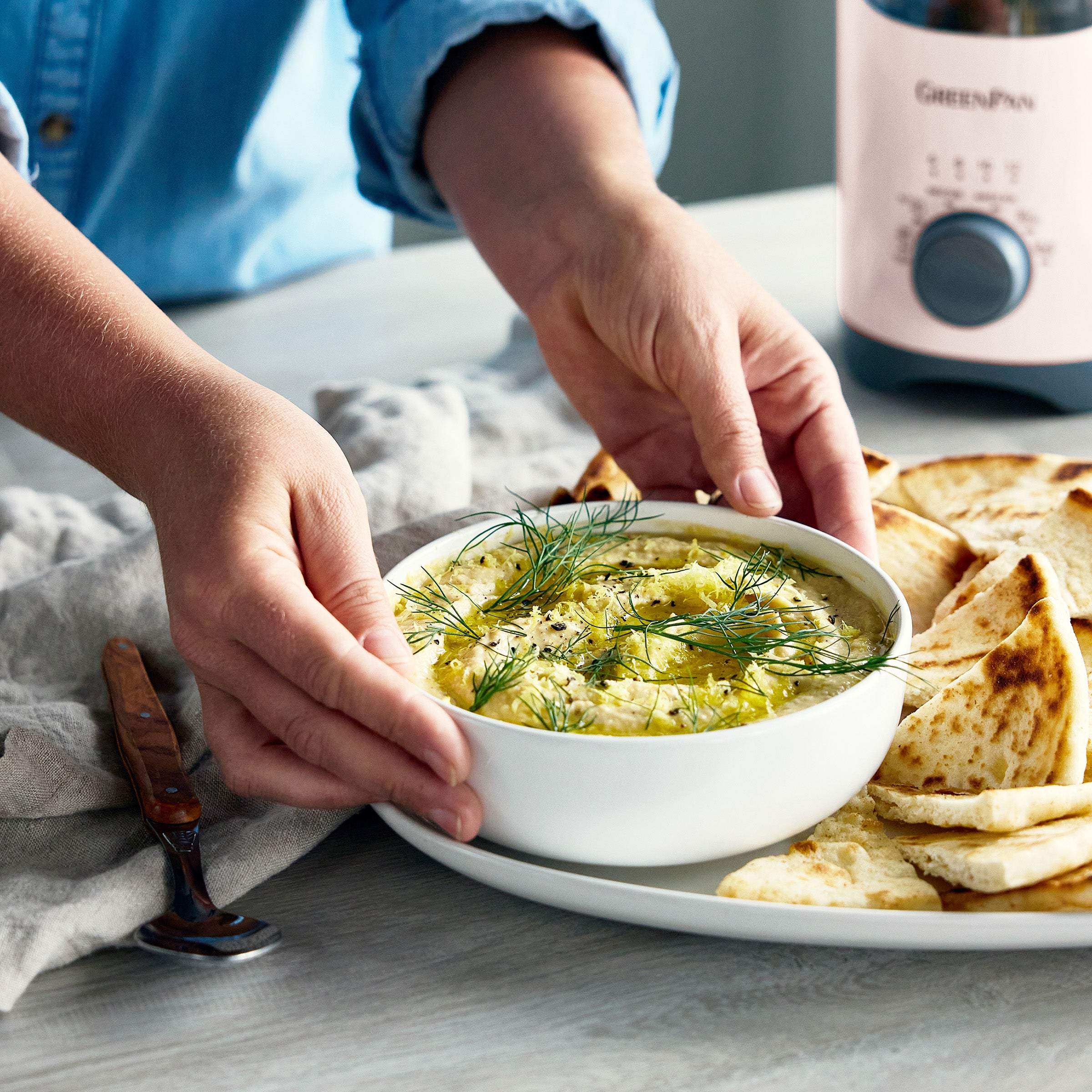 Person dipping bread into a bowl of hummus on a table with a kitchen appliance in the background.