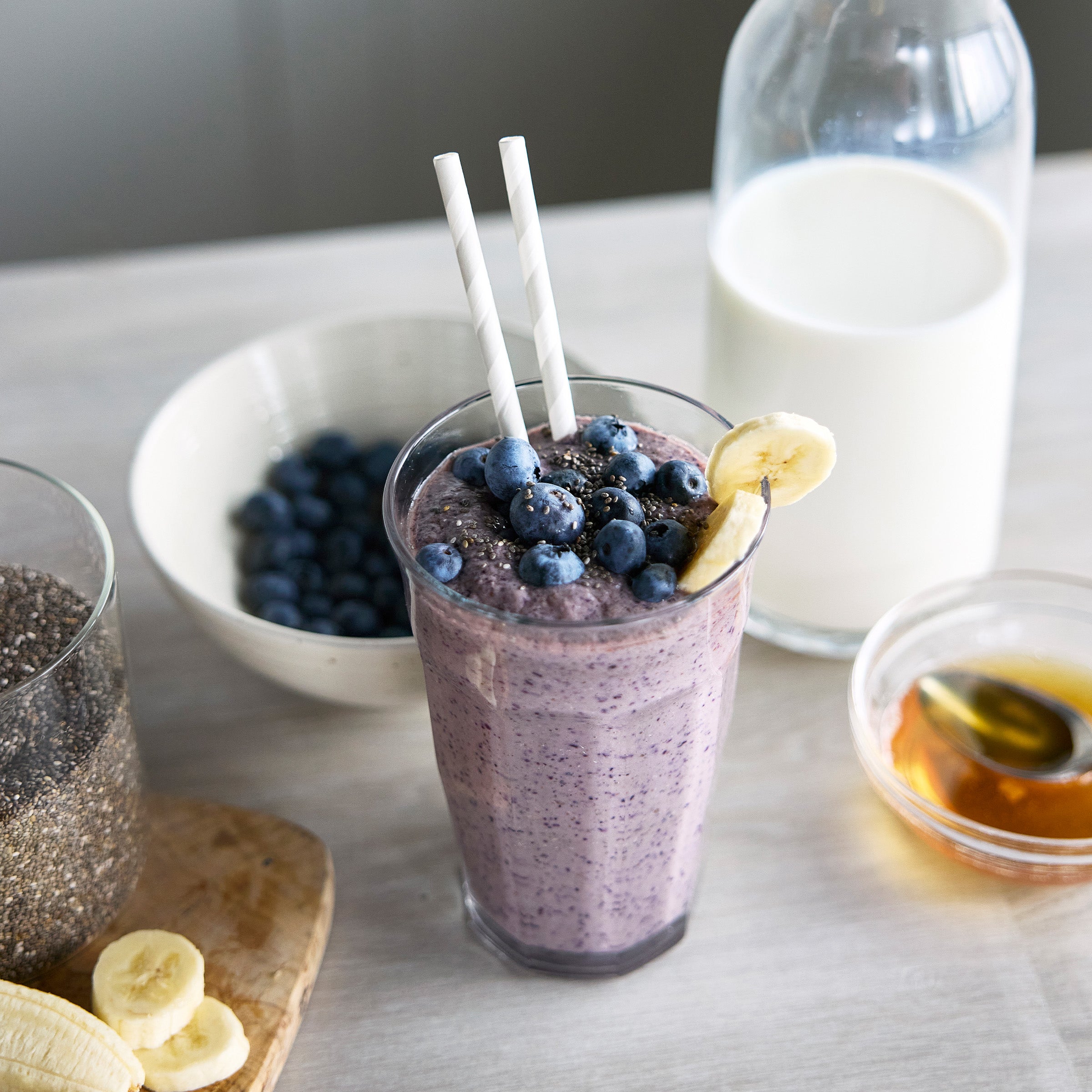 Blueberry smoothie with banana slices, a bowl of blueberries, and a jar of honey on a table.