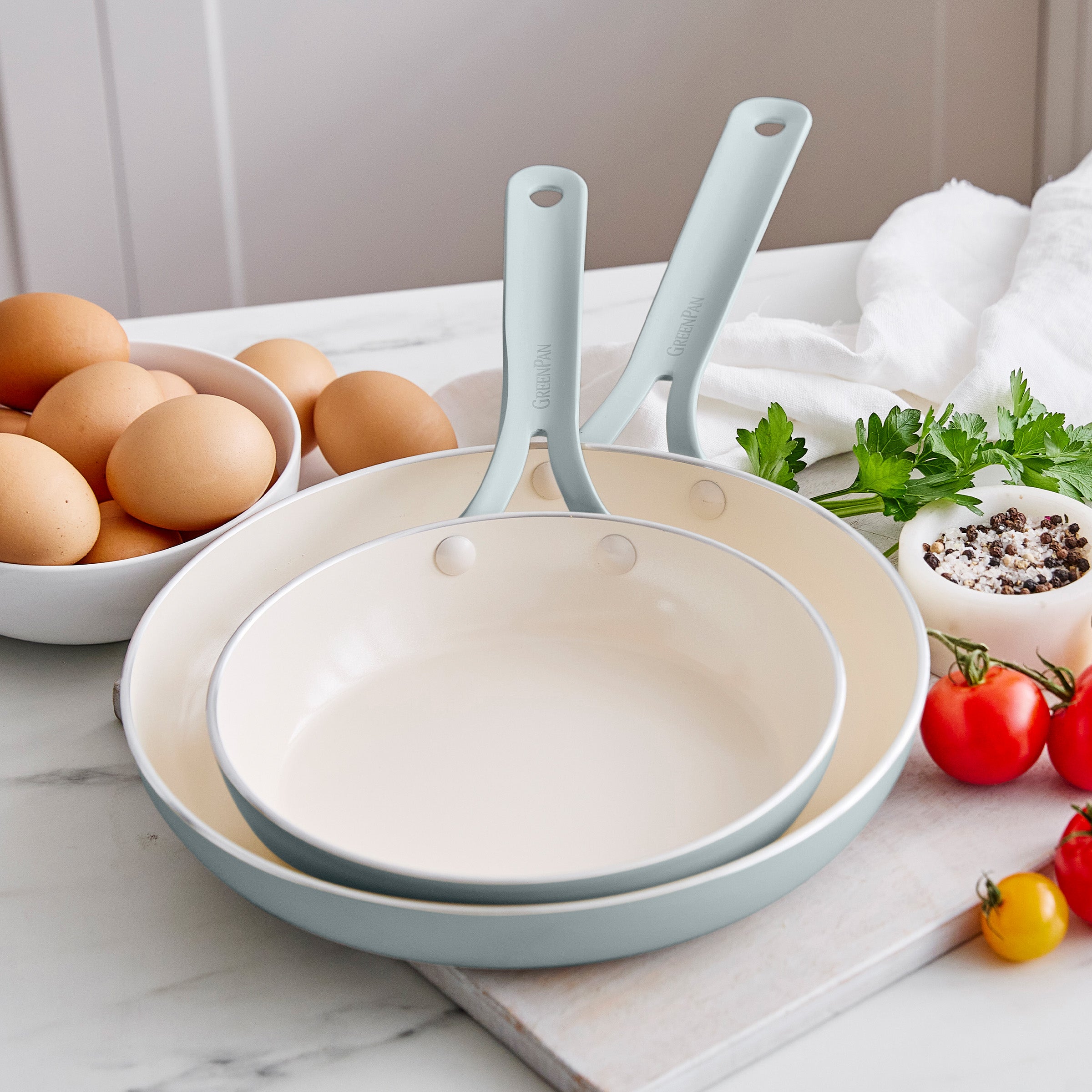 Set of frypans with handles on a kitchen counter with eggs and vegetables.