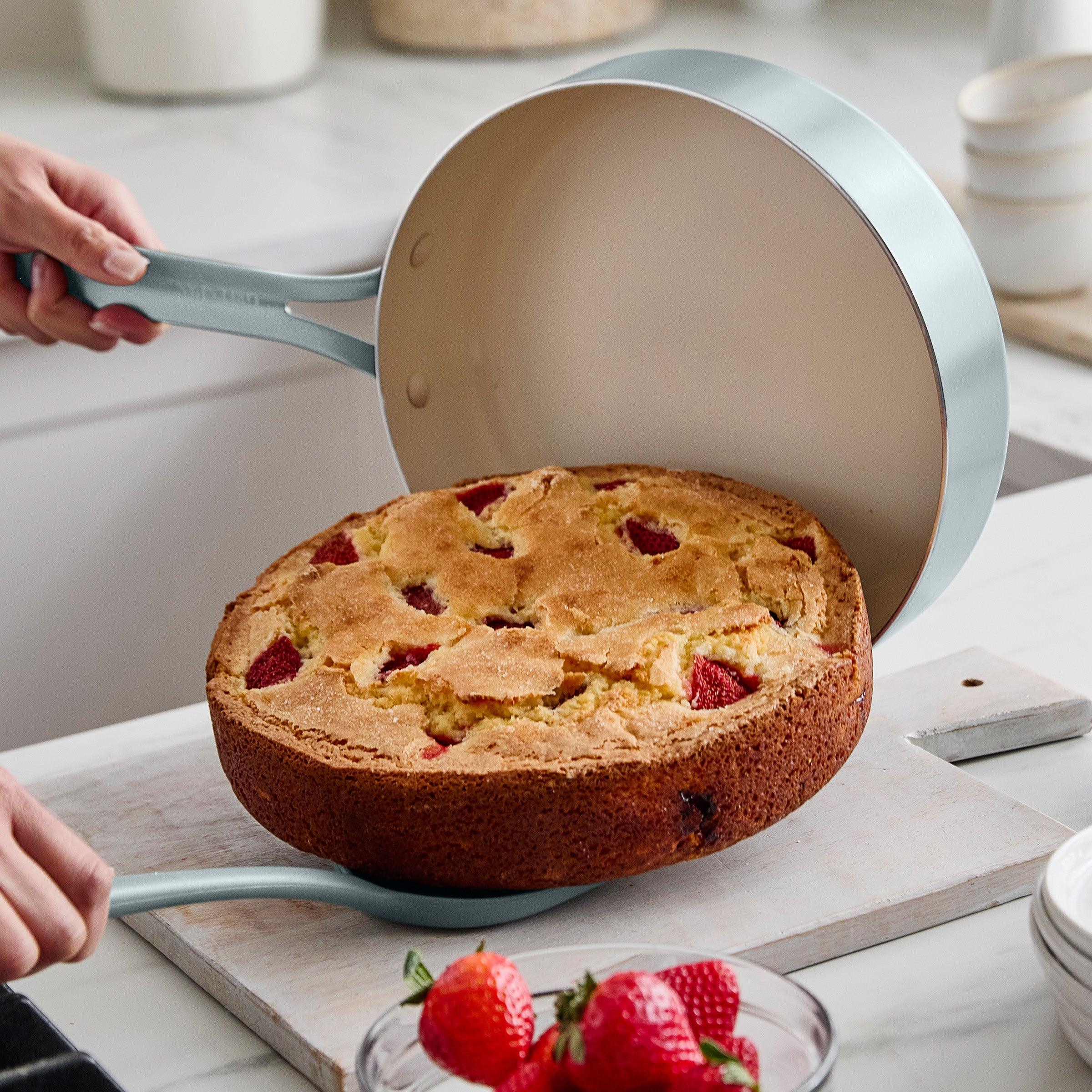 Person lifting a cake out of a round pan with strawberries on a kitchen counter.