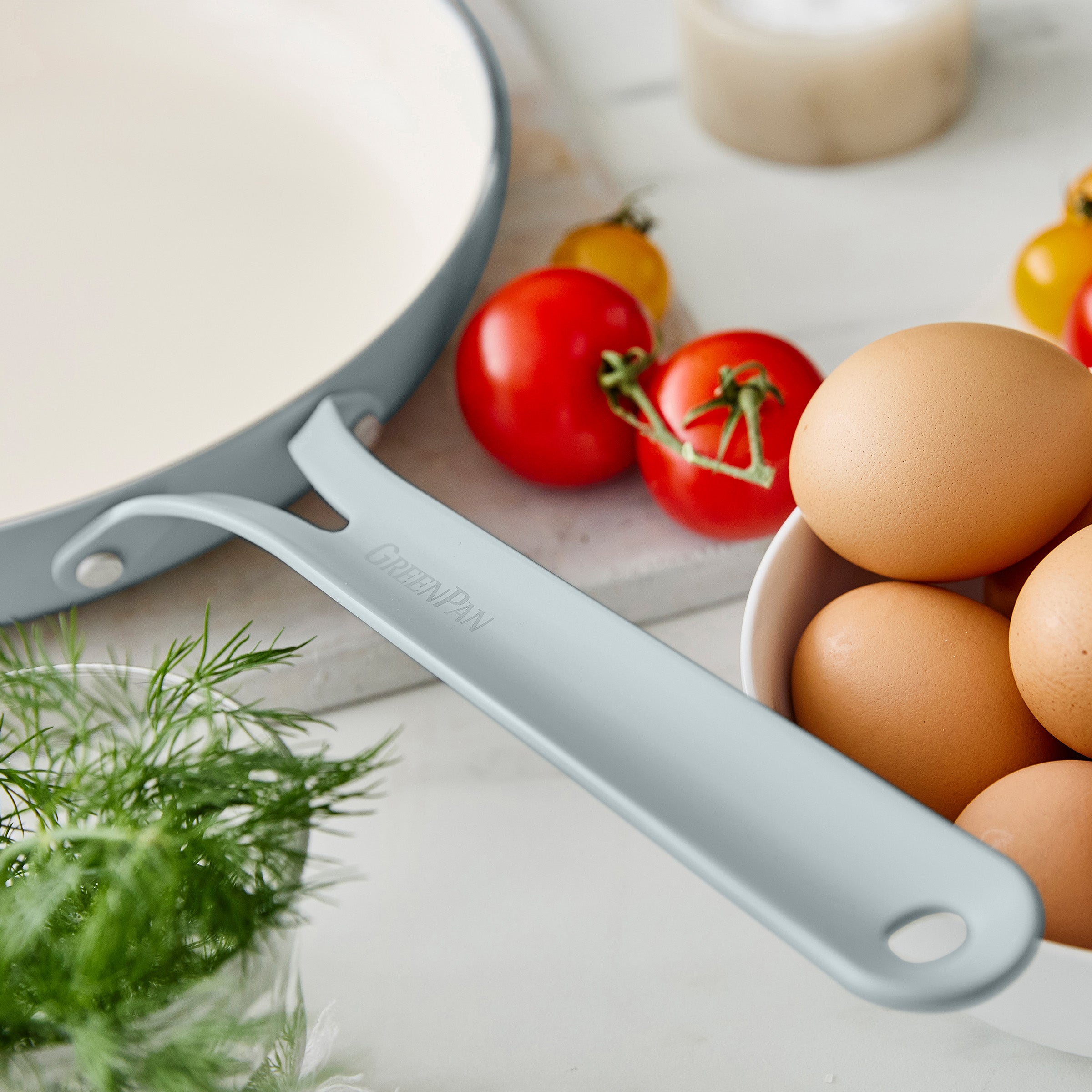 Light blue frypan handle on a white surface with eggs and tomatoes
