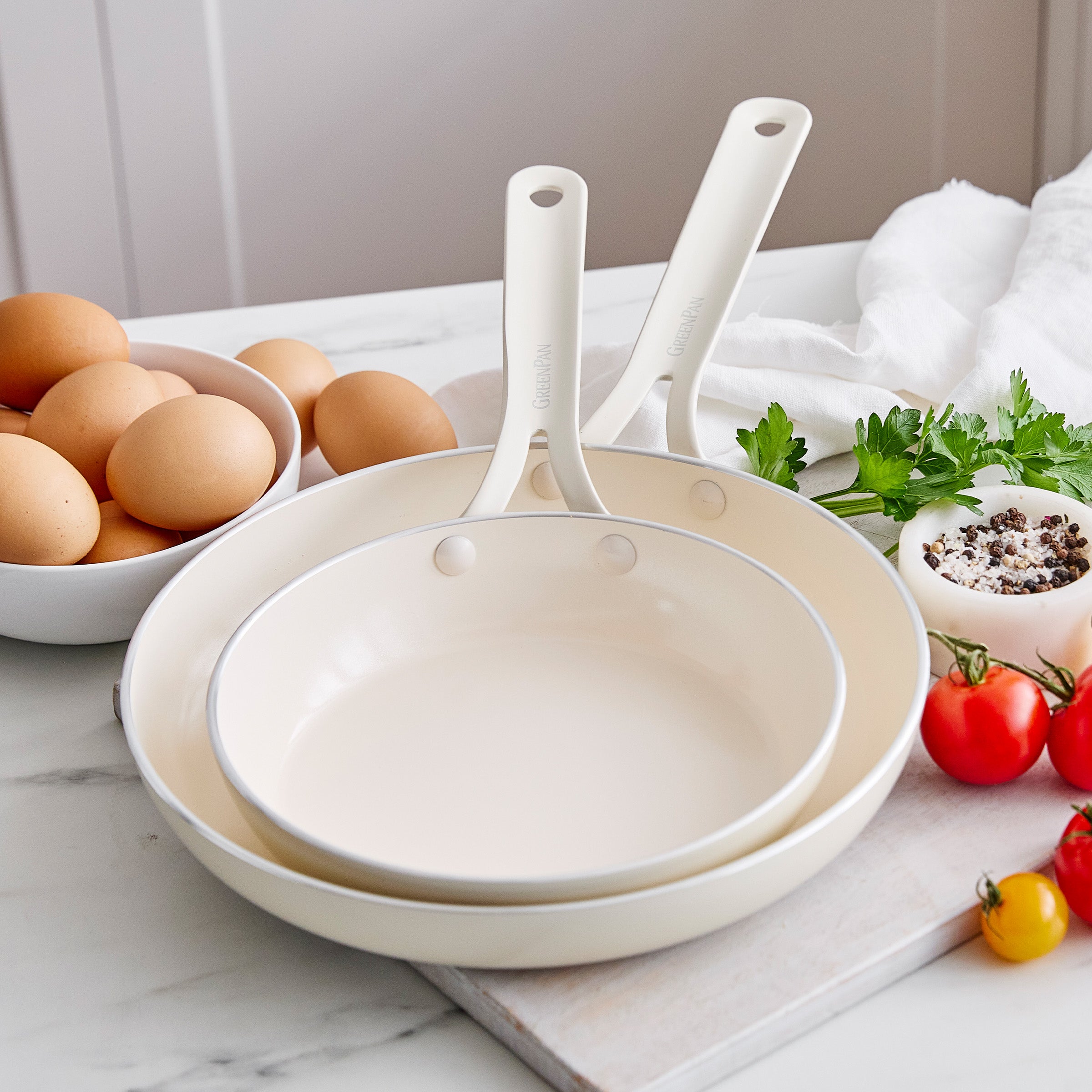 Set of white frypans on a marble surface with eggs and vegetables.
