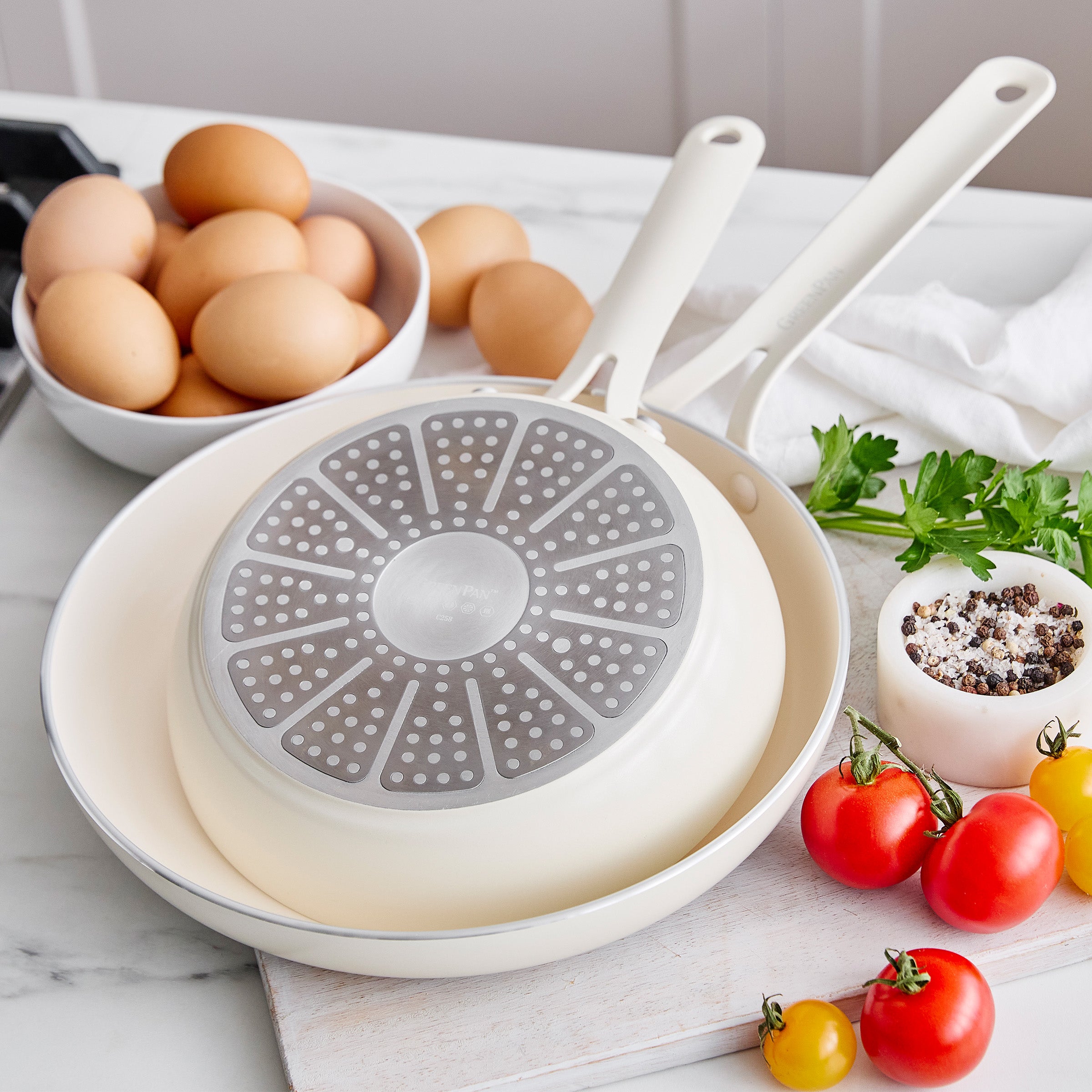 Cream frying pans on a kitchen counter with eggs and vegetables.