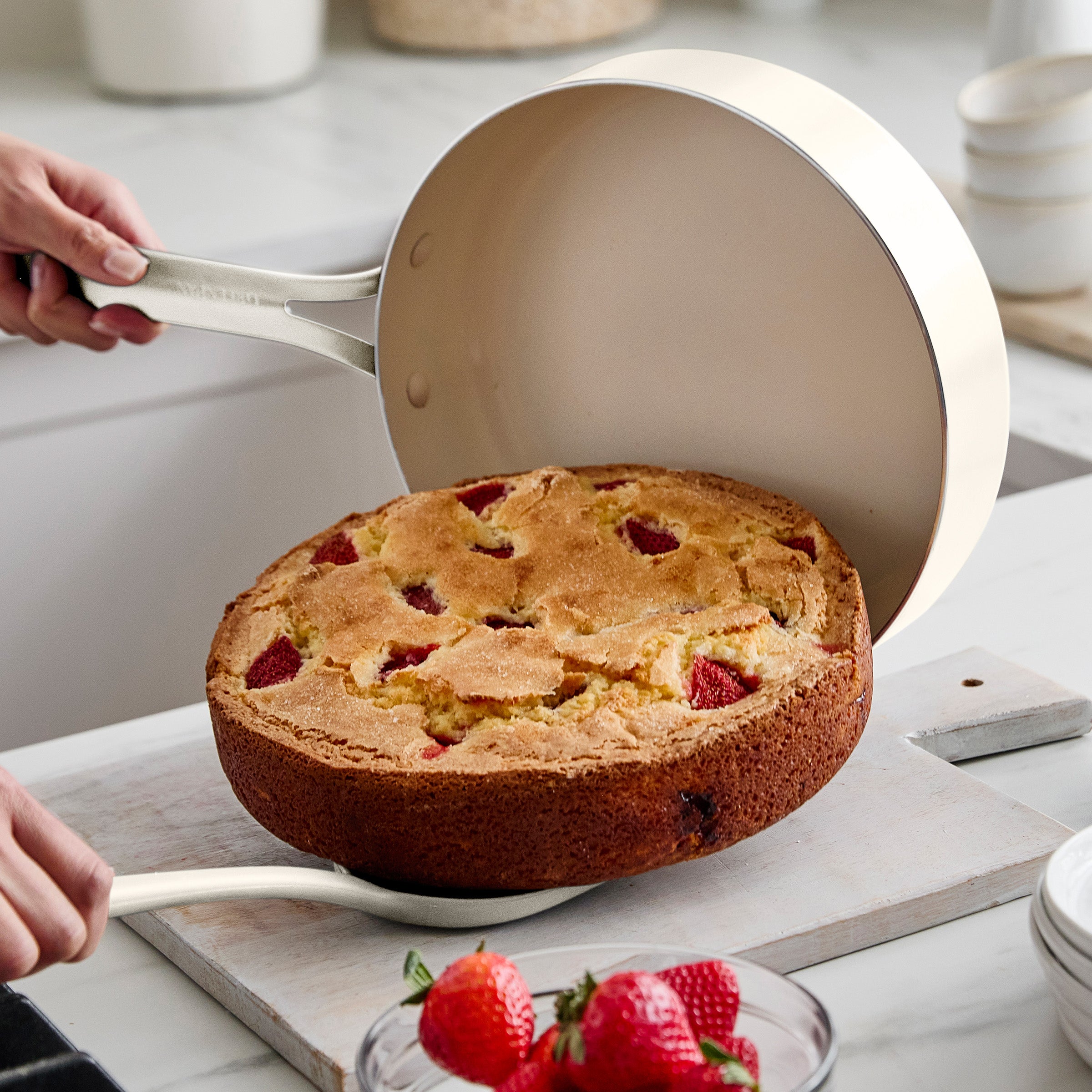 Person lifting a cake out of a round pan with strawberries on a kitchen counter.