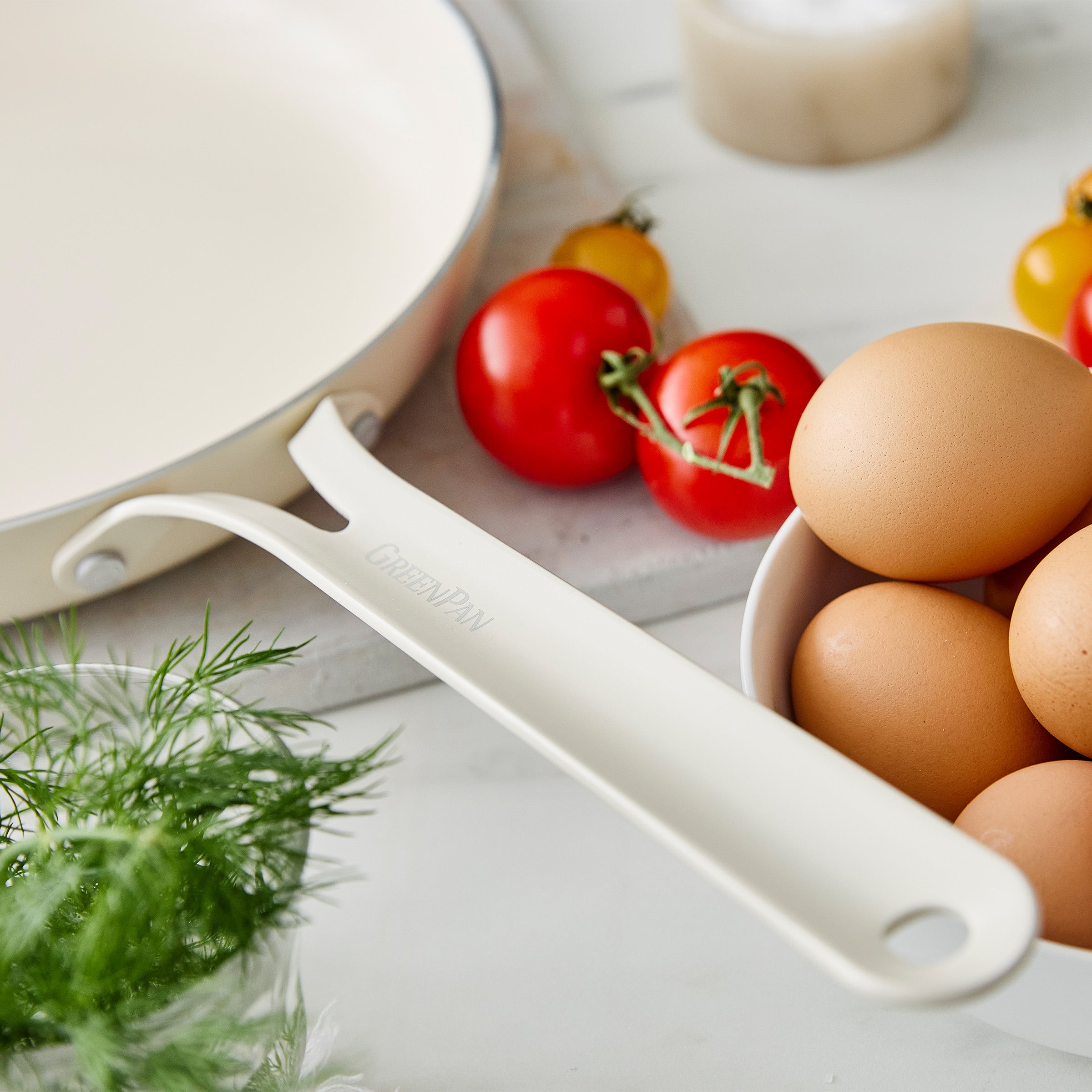 Cream frypan handle on a surface with eggs, tomatoes, and herbs.