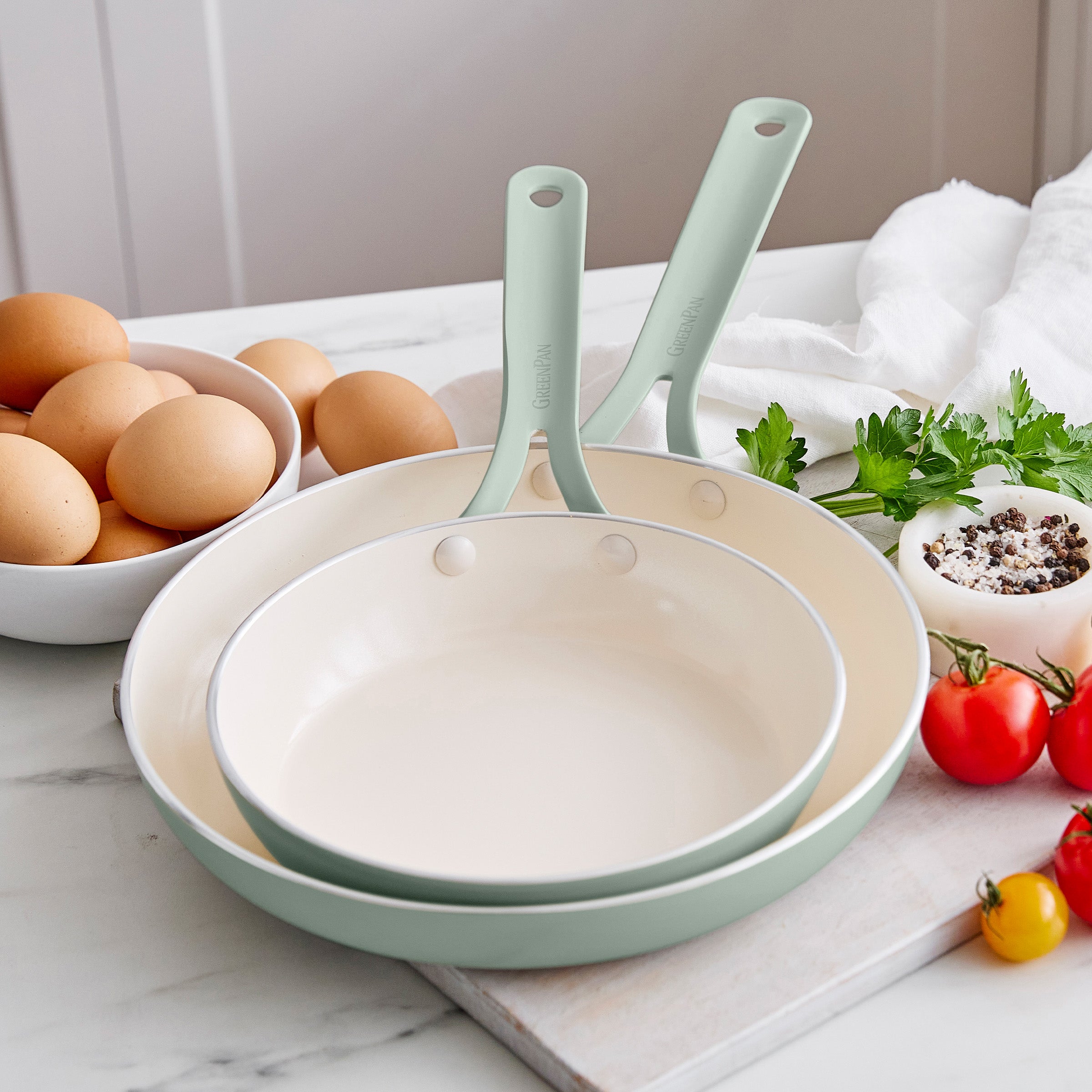 Set of frypans with green handles on a kitchen counter with eggs and vegetables.
