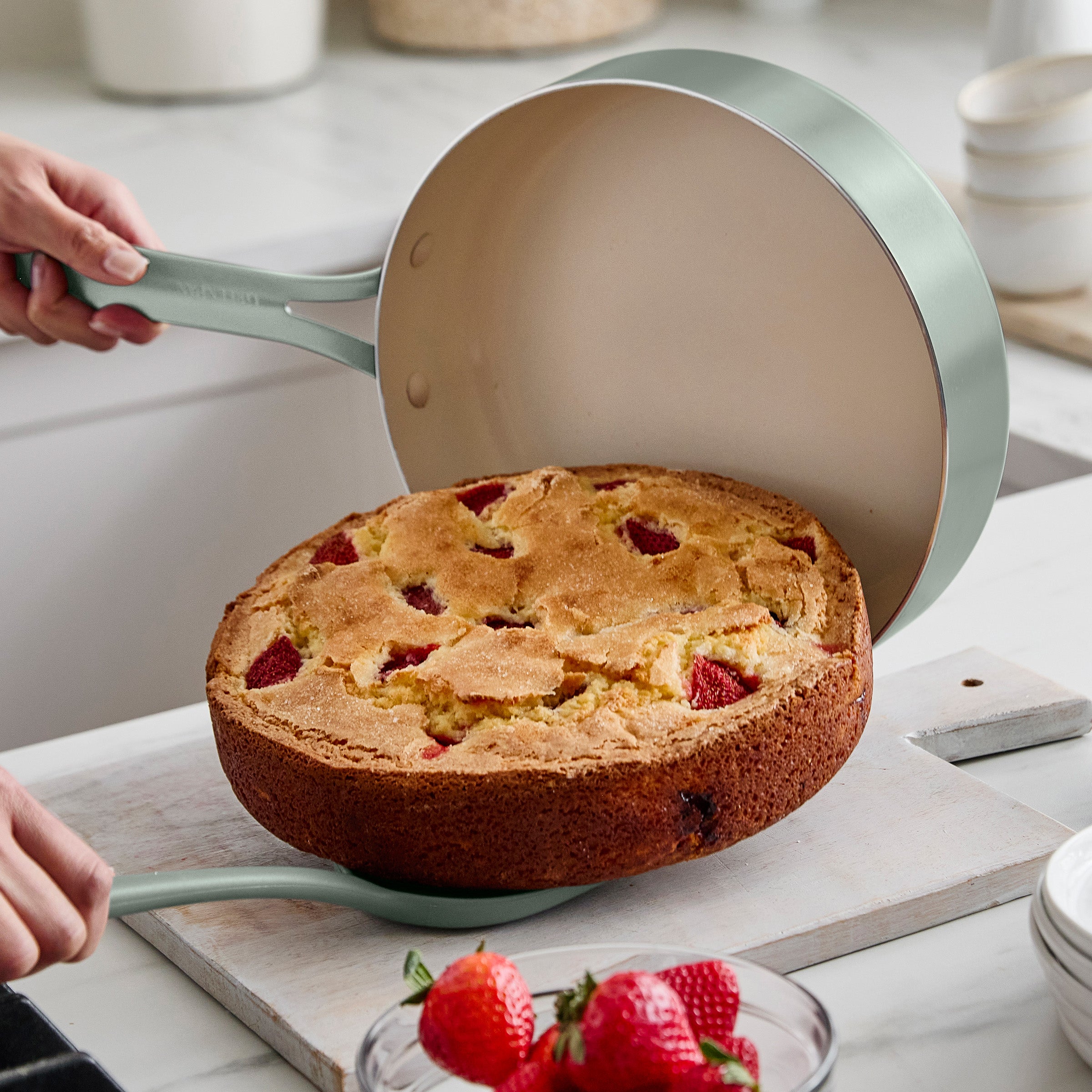 Person lifting a cake out of a round pan with strawberries on a kitchen counter.