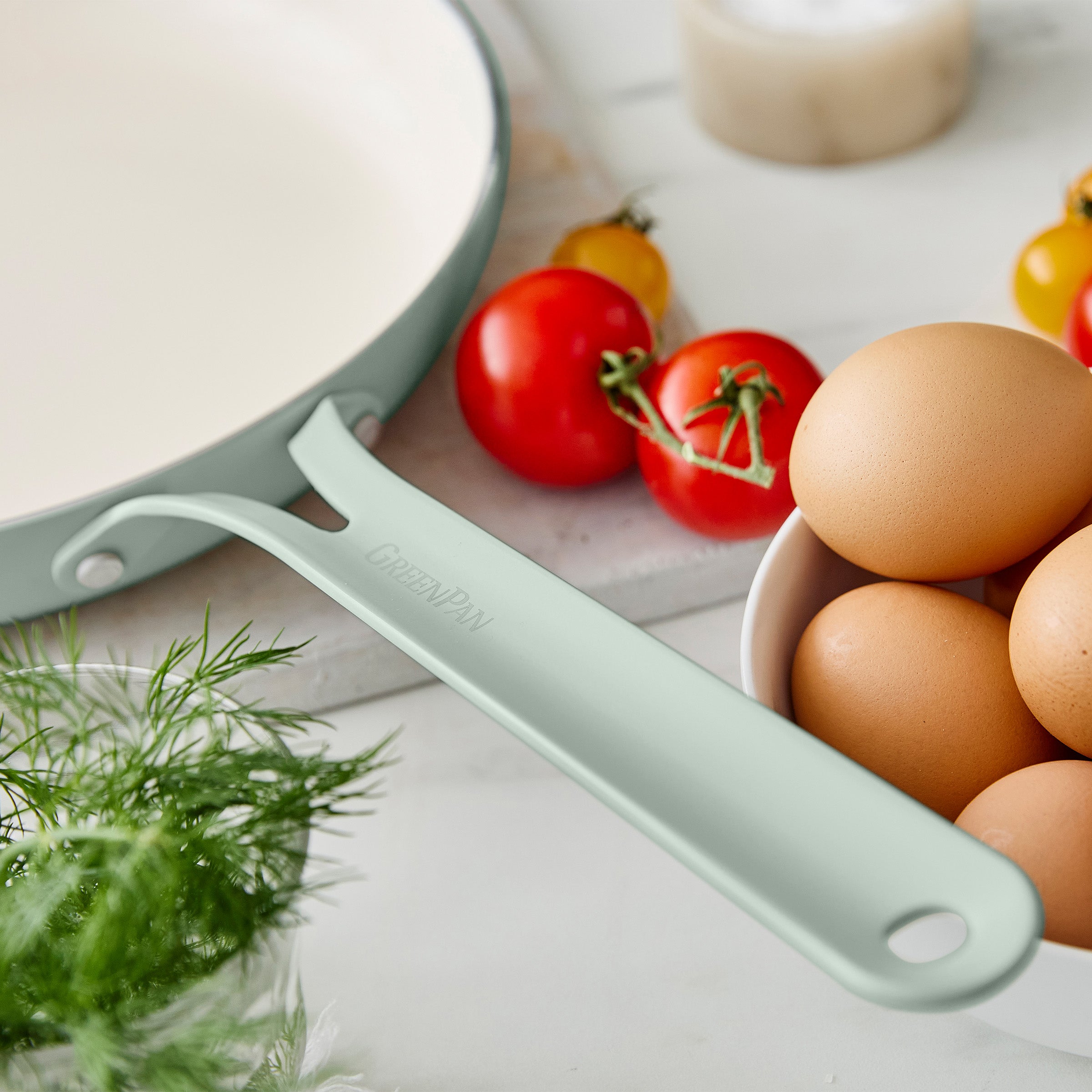 Green frypan handle on a white surface with tomatoes and eggs