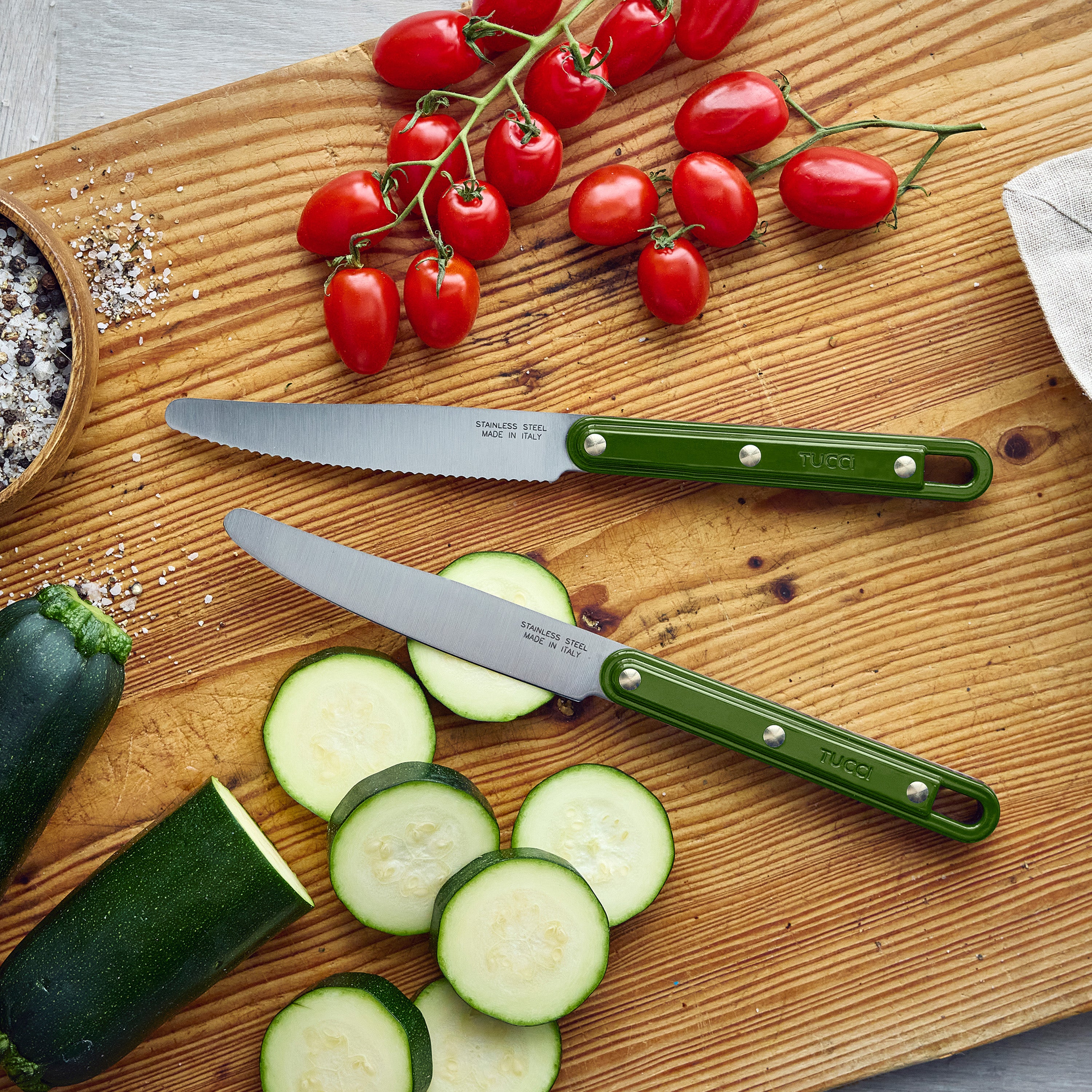 Two knives on a wooden cutting board with sliced zucchini and tomatoes, featuring the brand 'Tucci'