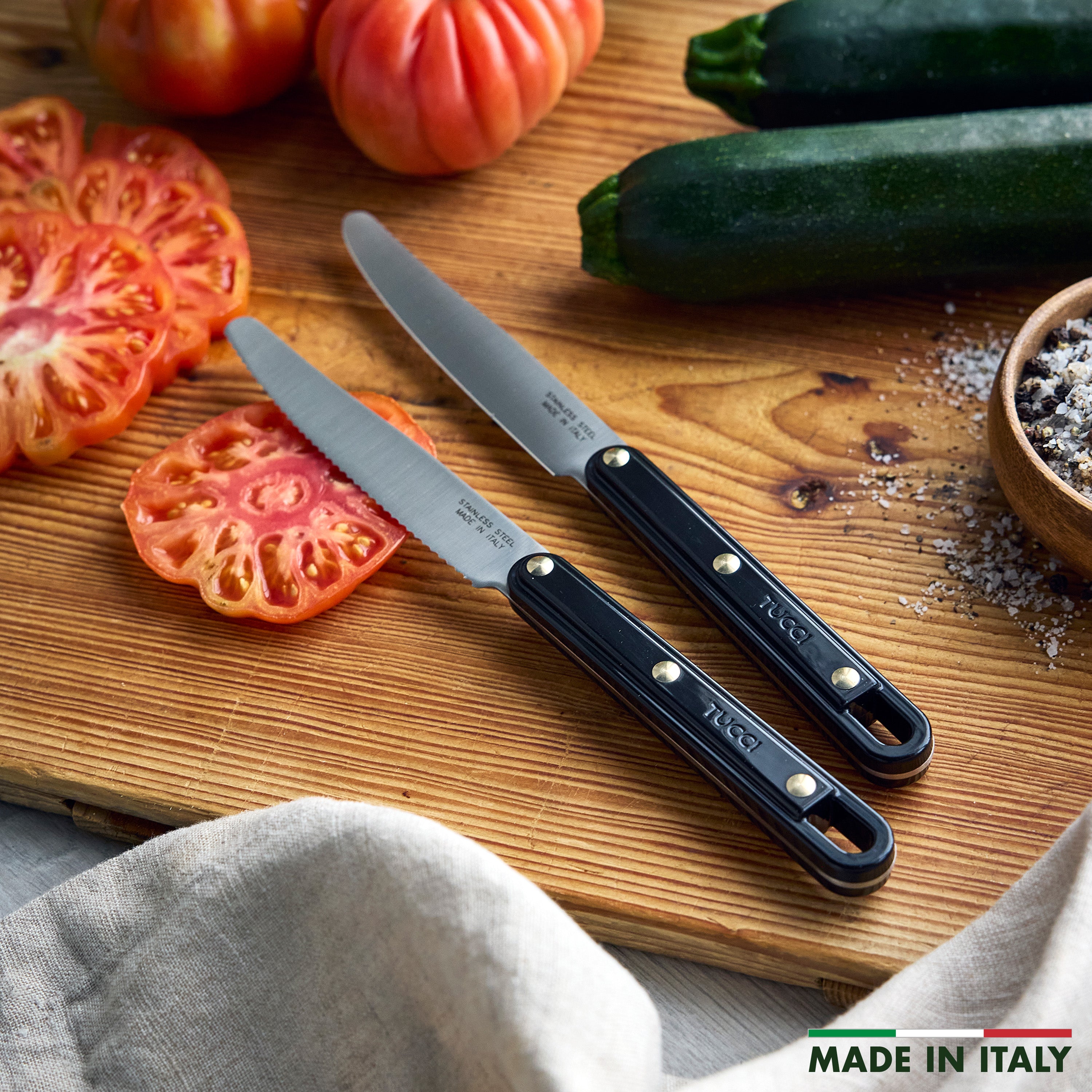 Two knives on a wooden cutting board with sliced zucchini and tomatoes, featuring the brand 'Tucci'