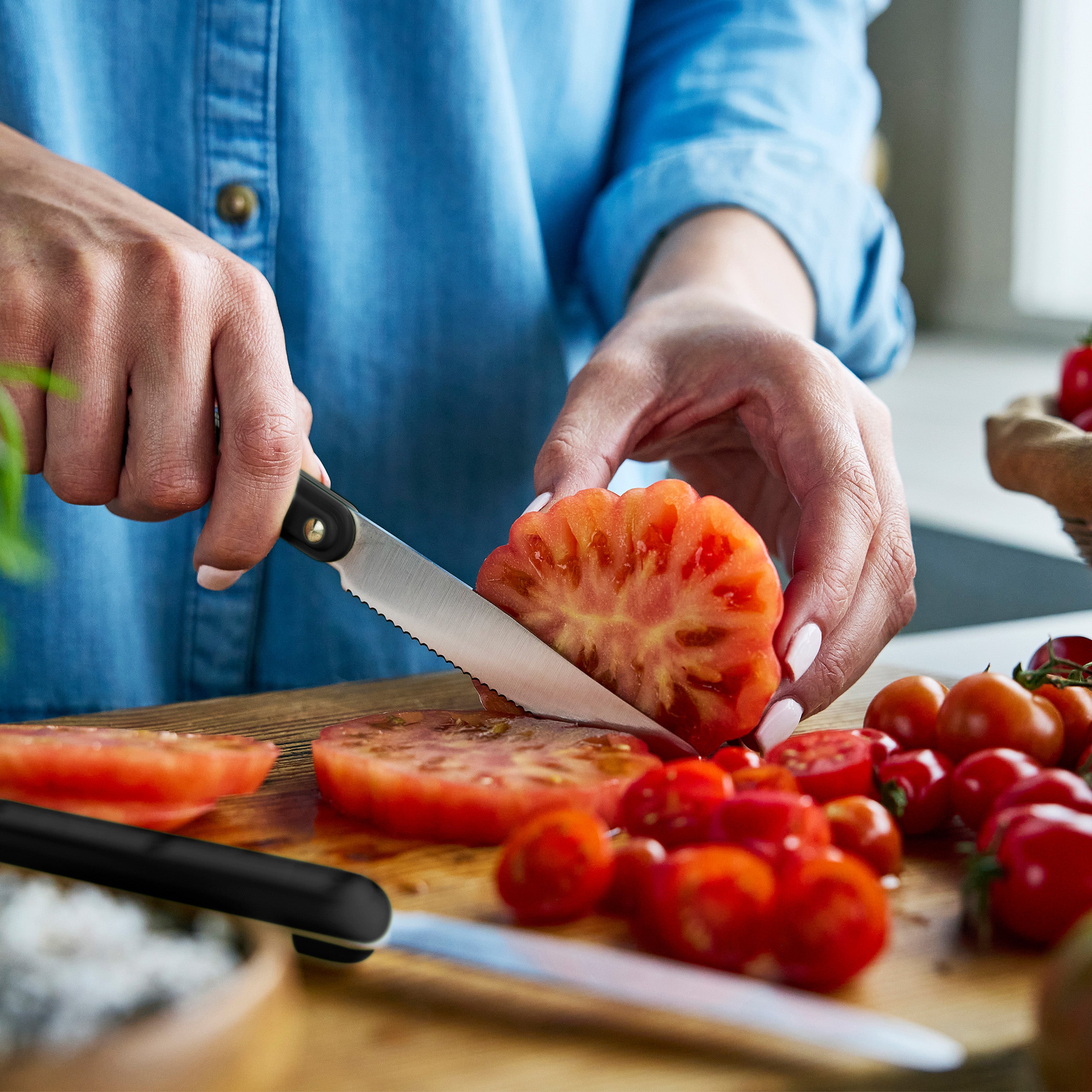 Person slicing tomato on a wooden cutting board with a knife.