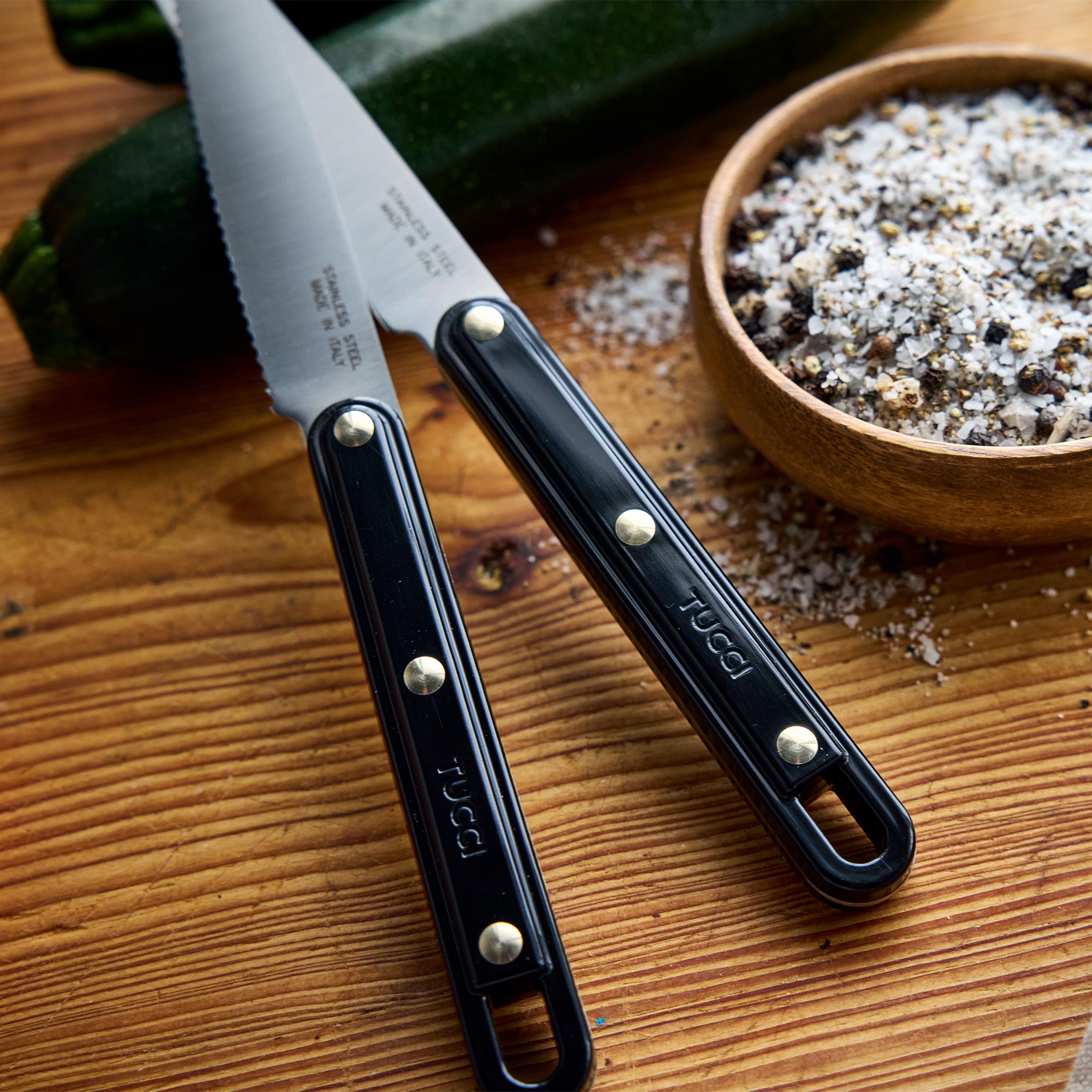 Two knives with black handles on a wooden cutting board with zucchini and seasoning