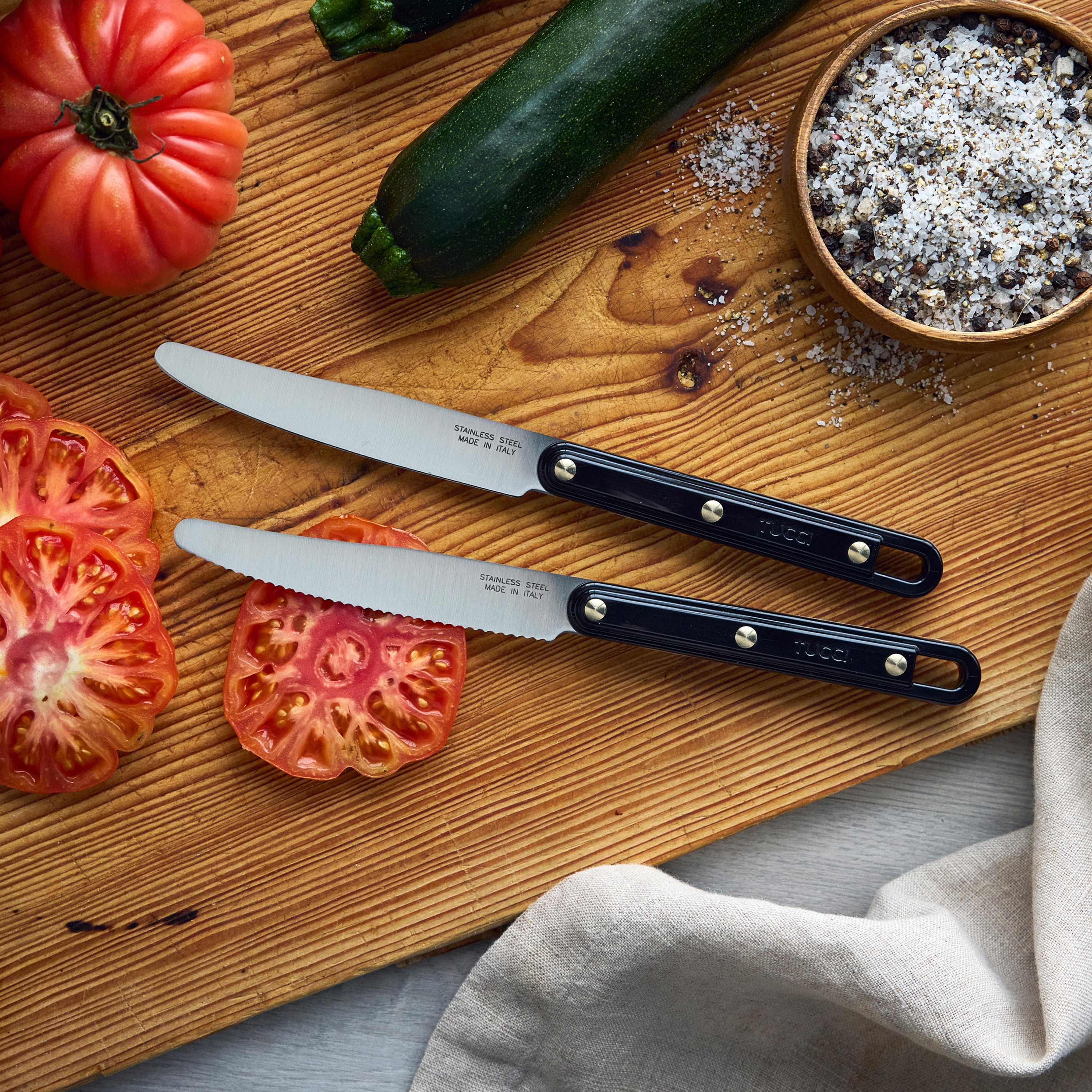Two knives on a wooden cutting board with zucchini and tomatoes, featuring the brand 'Tucci'