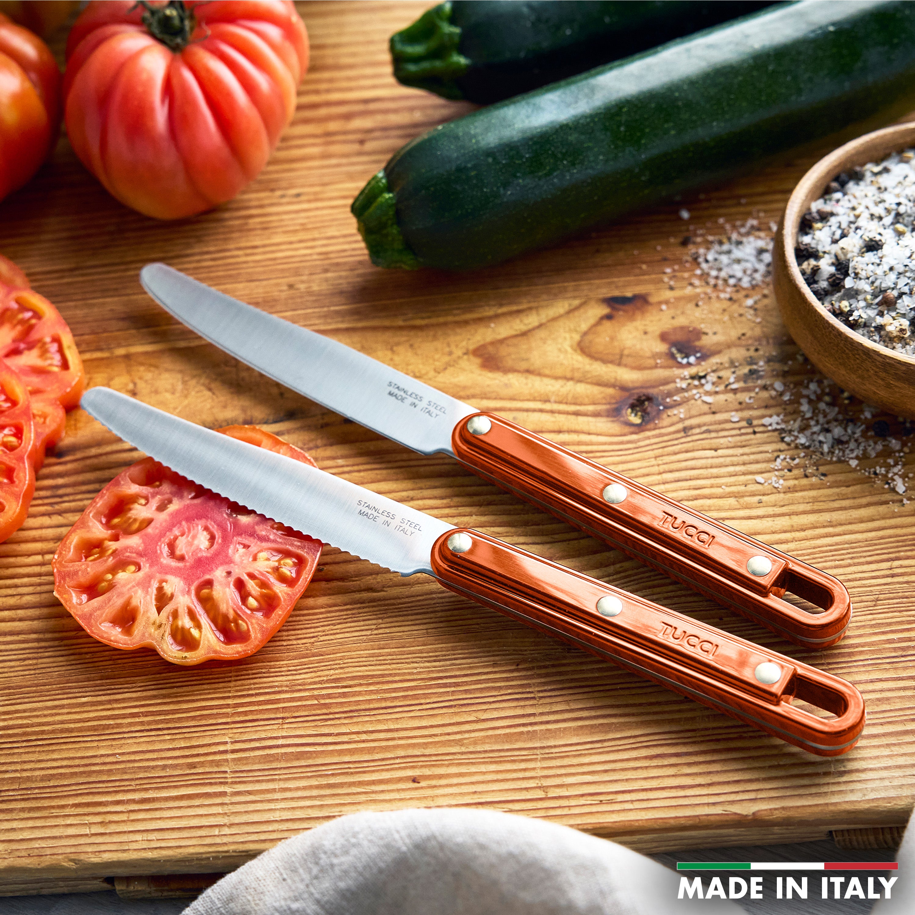 Two knives on a wooden cutting board with sliced zucchini and tomatoes, featuring the brand 'Tucci'