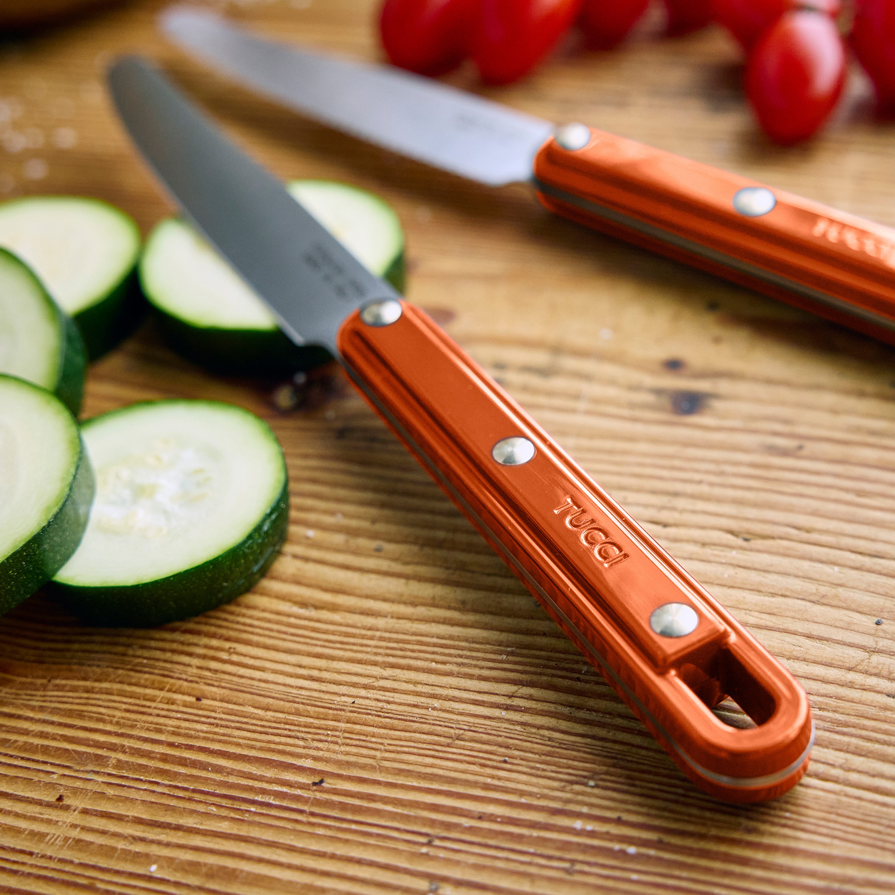Two knives with orange handles on a wooden cutting board with zucchini and tomatoes