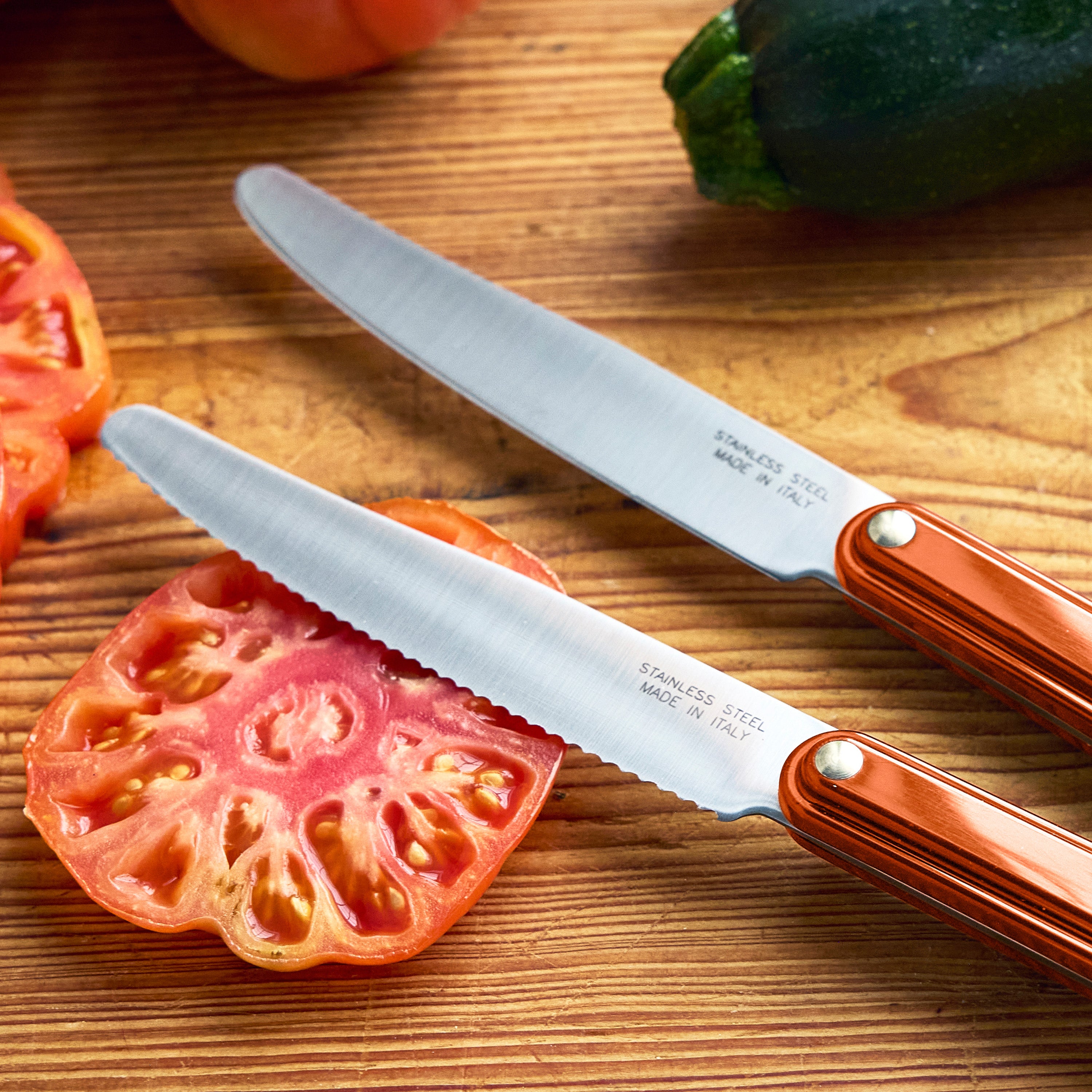 Two knives on a wooden cutting board with sliced zucchini and tomatoes, featuring the brand 'Tucci'