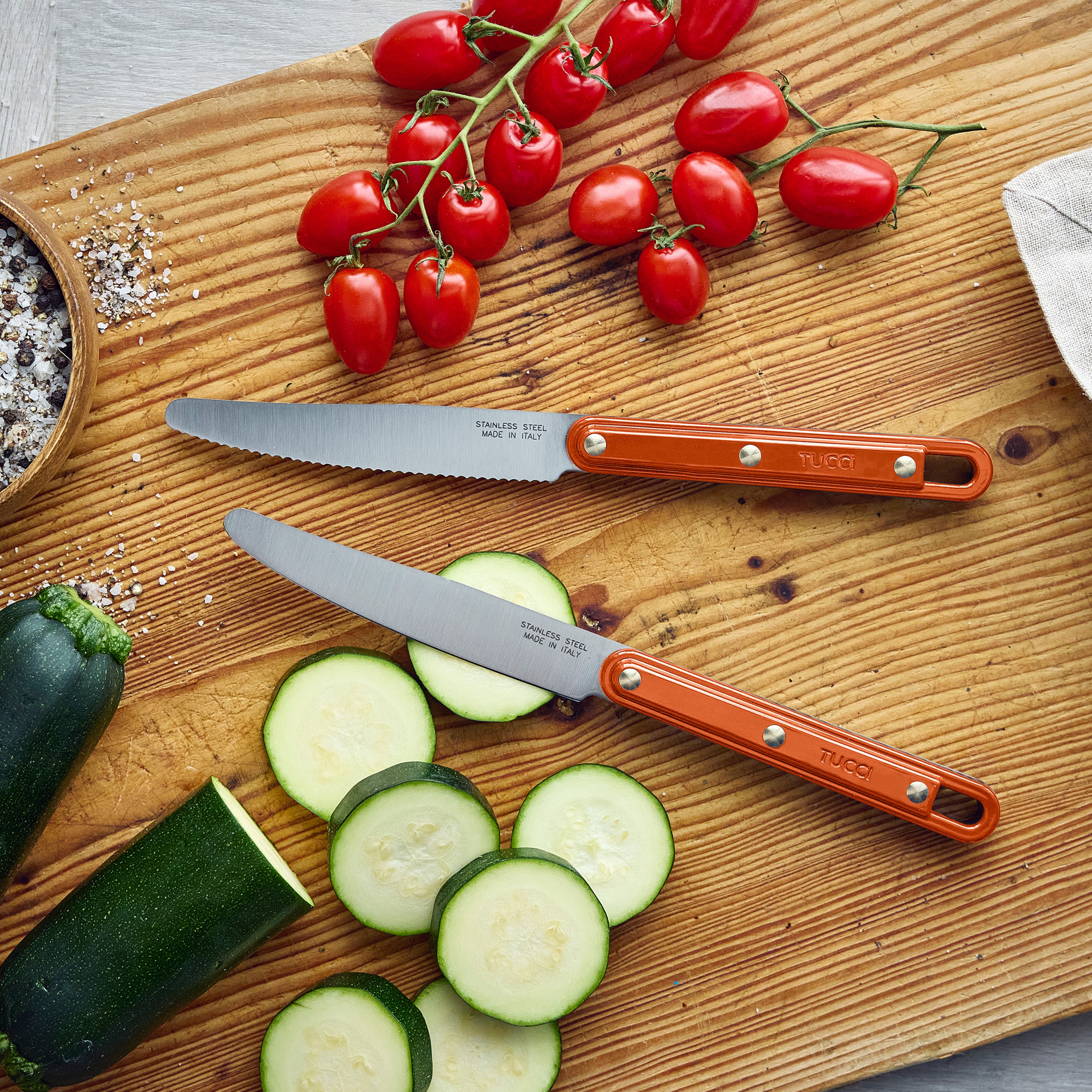 Two orange-handled knives on a wooden cutting board with sliced zucchini and cherry tomatoes.