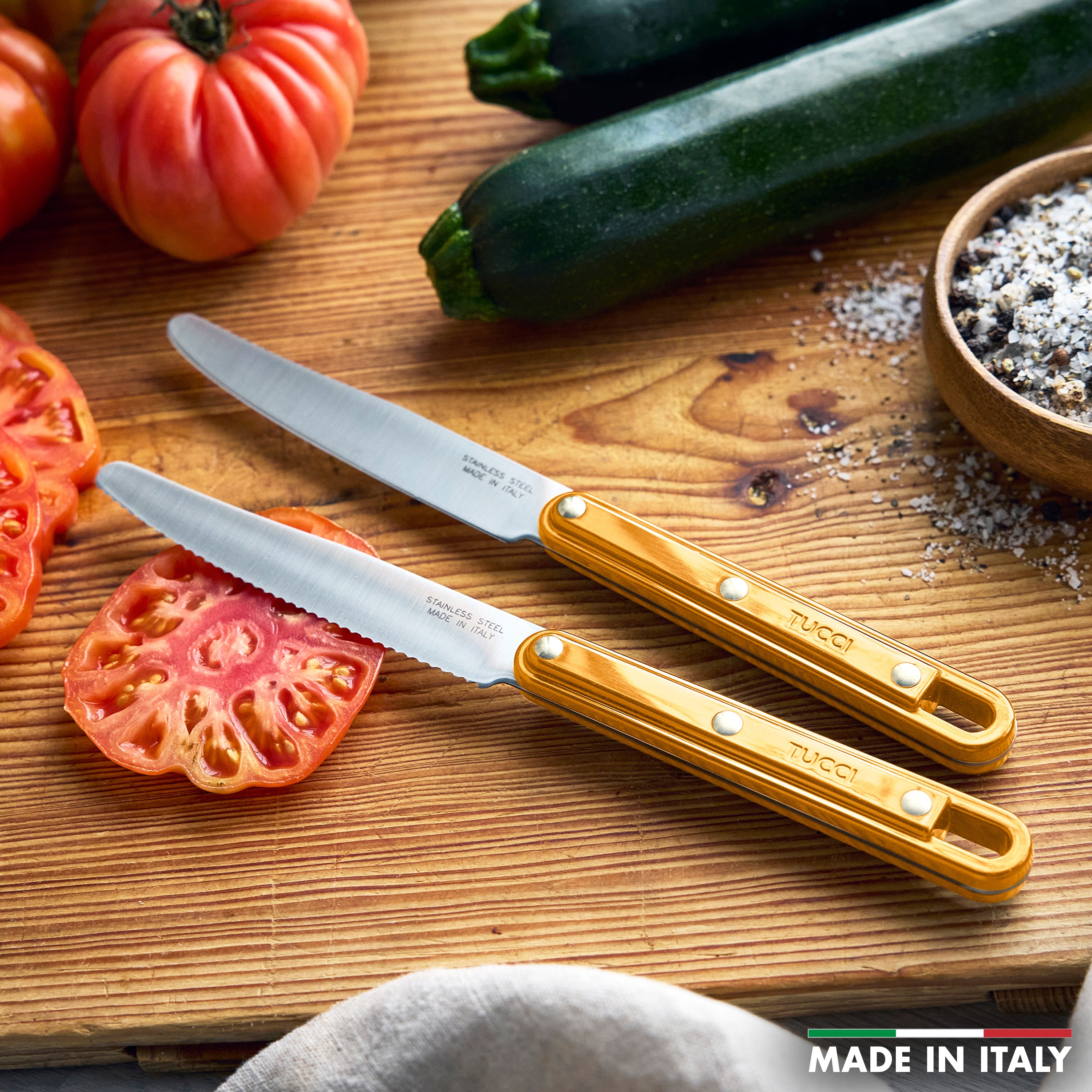 Two knives on a wooden cutting board with sliced zucchini and tomatoes, featuring the brand 'Tucci'