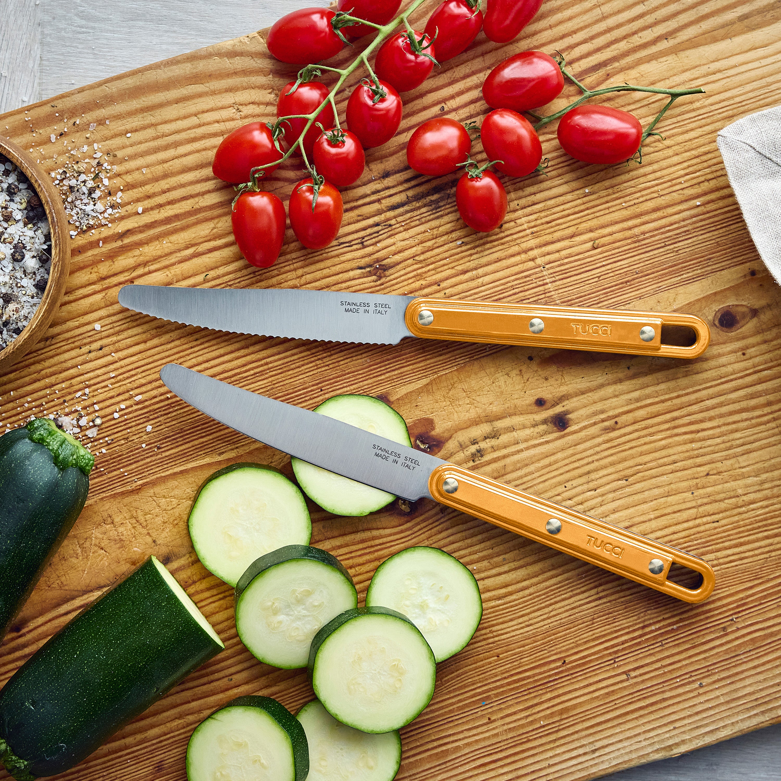 Two yellow-handled knives on a wooden cutting board with sliced zucchini and cherry tomatoes.