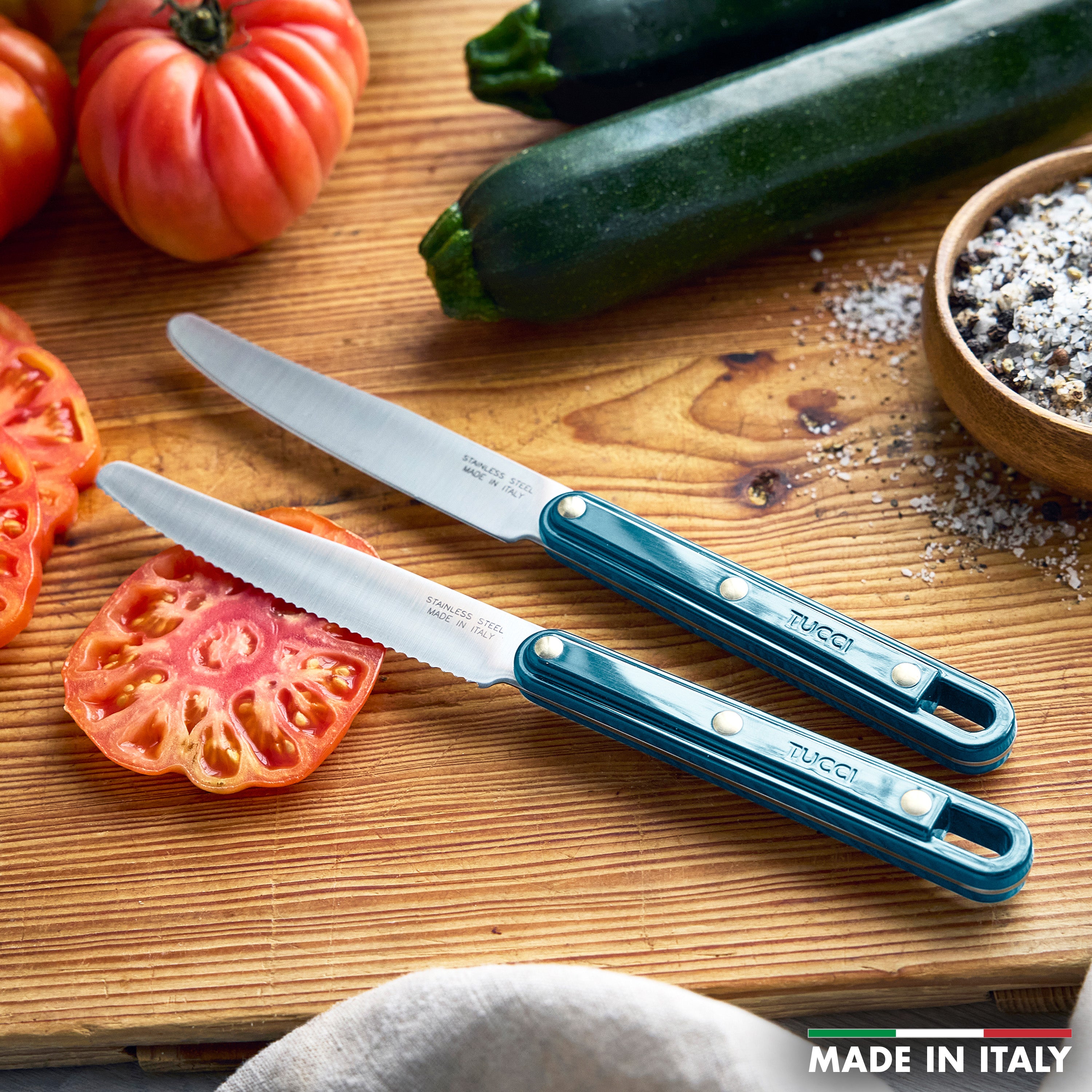 Two knives on a wooden cutting board with sliced zucchini and tomatoes, featuring the brand 'Tucci'