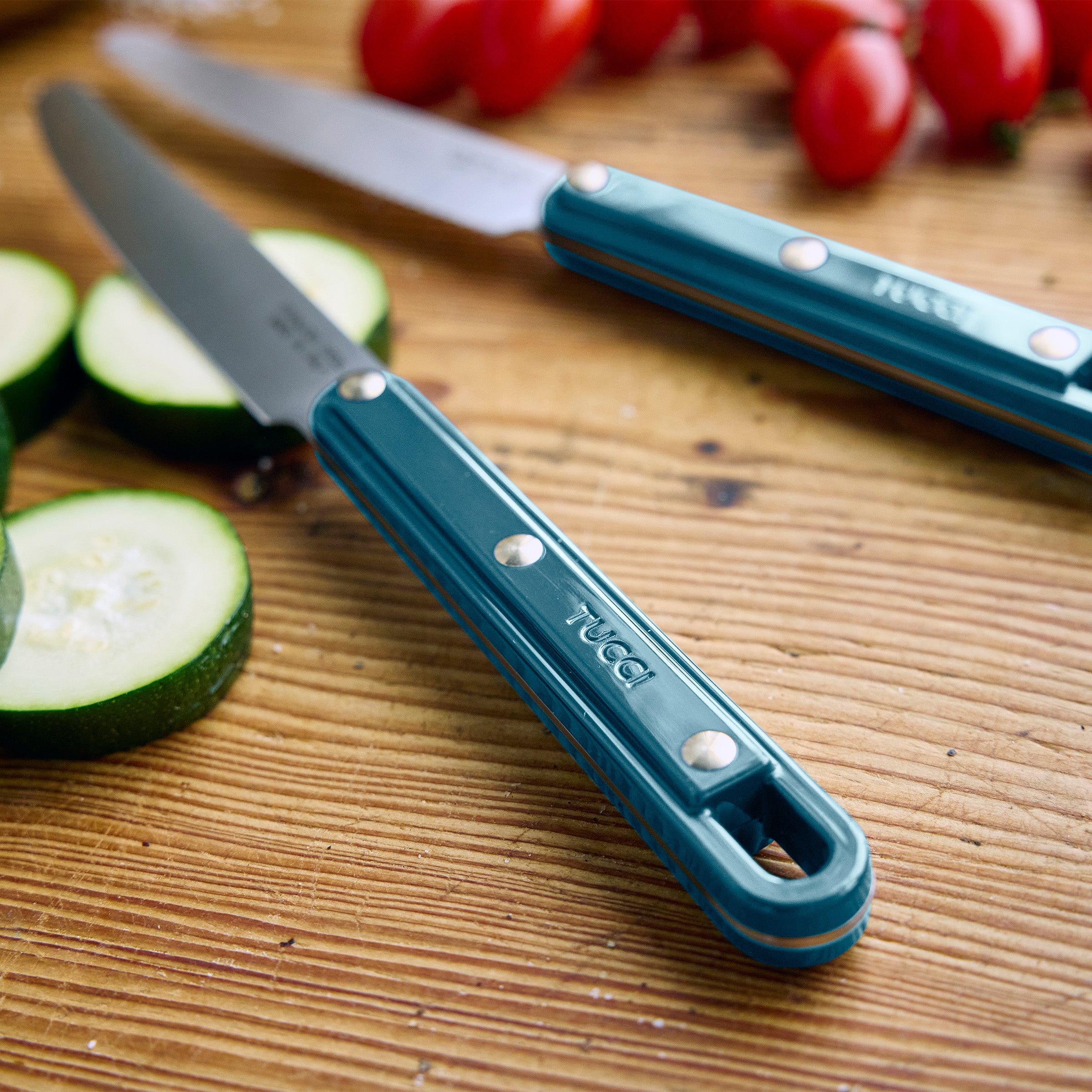 Two knives with teal handles on a wooden cutting board with zucchini and tomatoes