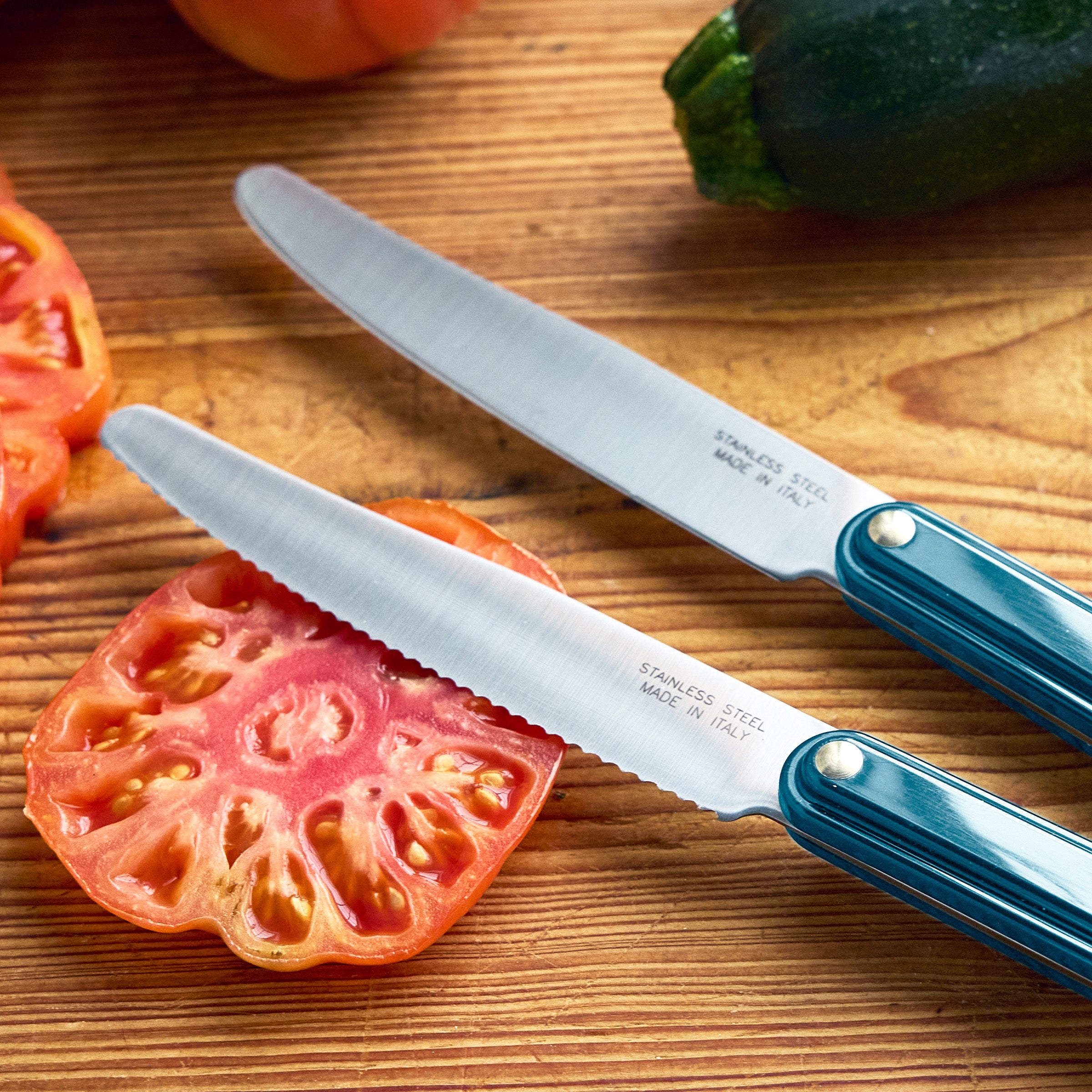 Two knives on a wooden cutting board with sliced zucchini and tomatoes, featuring the brand 'Tucci'