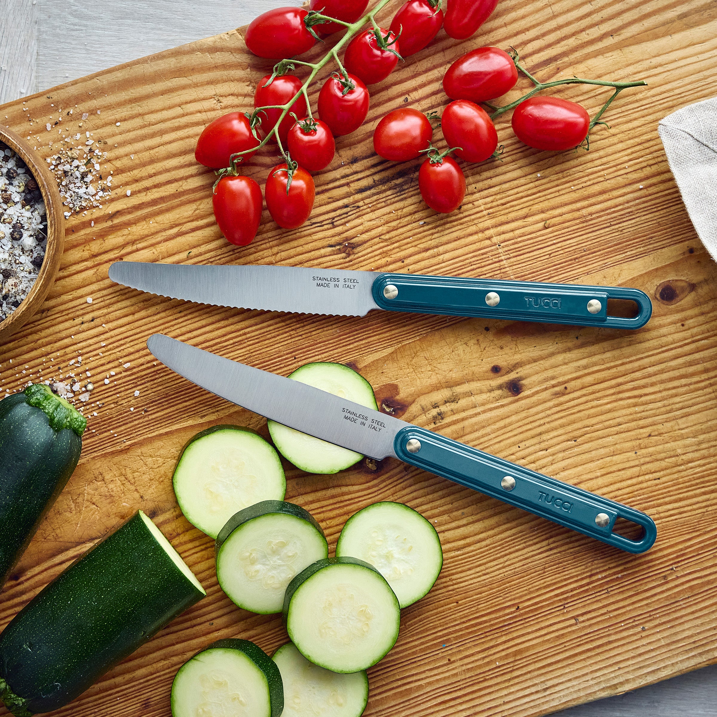 Two teal-handled knives on a wooden cutting board with sliced zucchini and cherry tomatoes.