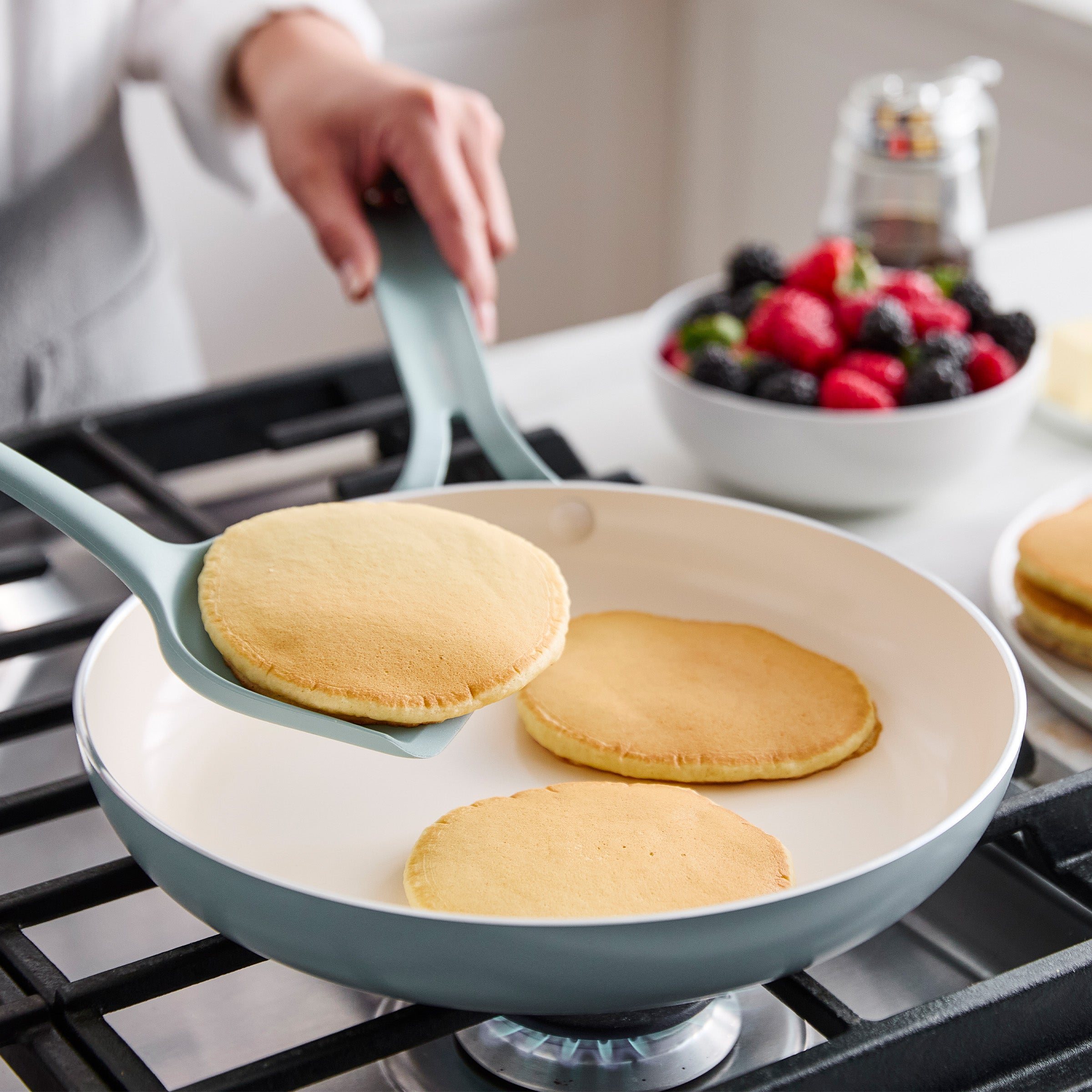 Person lifting a pancake from a frying pan with a turner, surrounded by a bowl of berries and other food items.