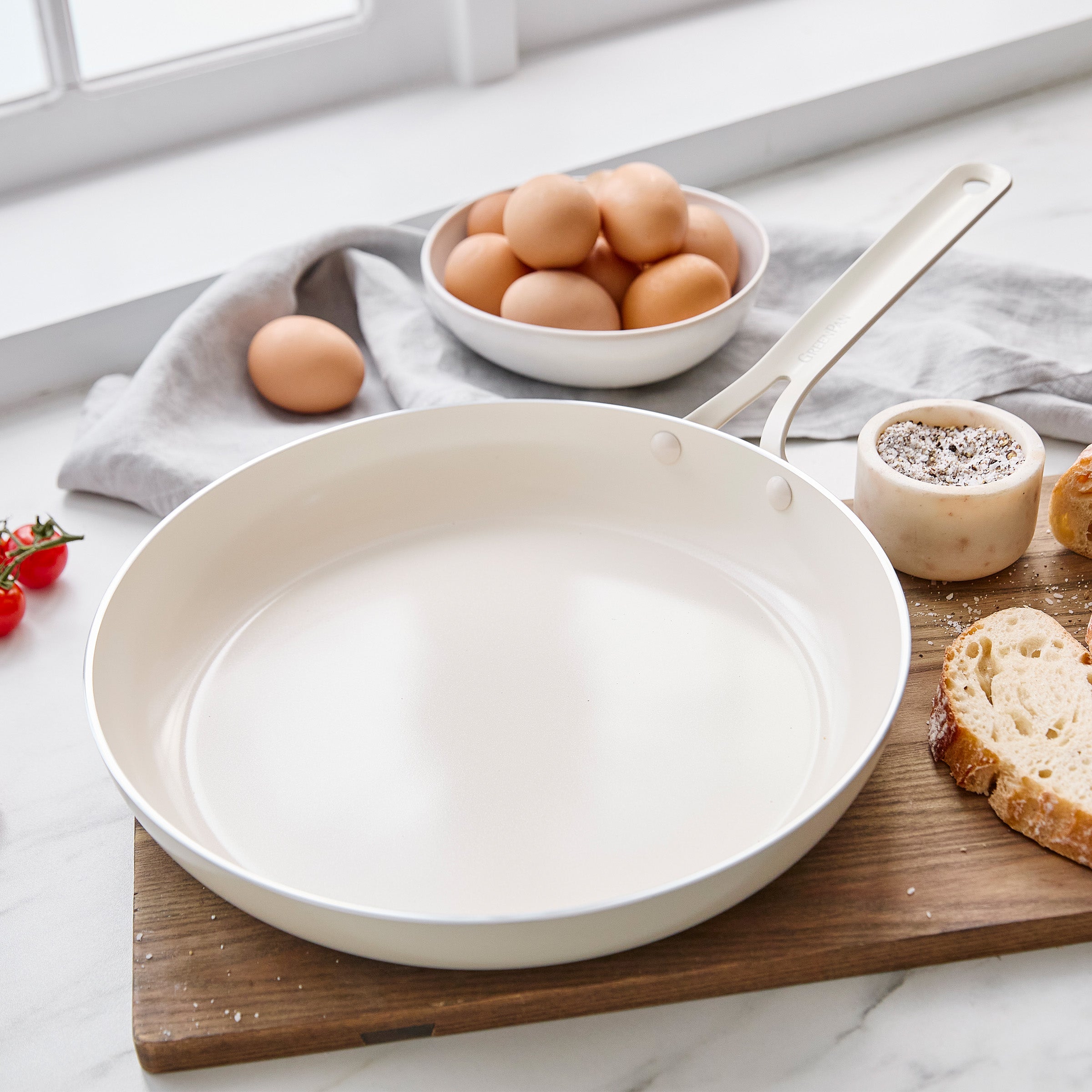 White frying pan on a wooden cutting board with eggs and bread in the background