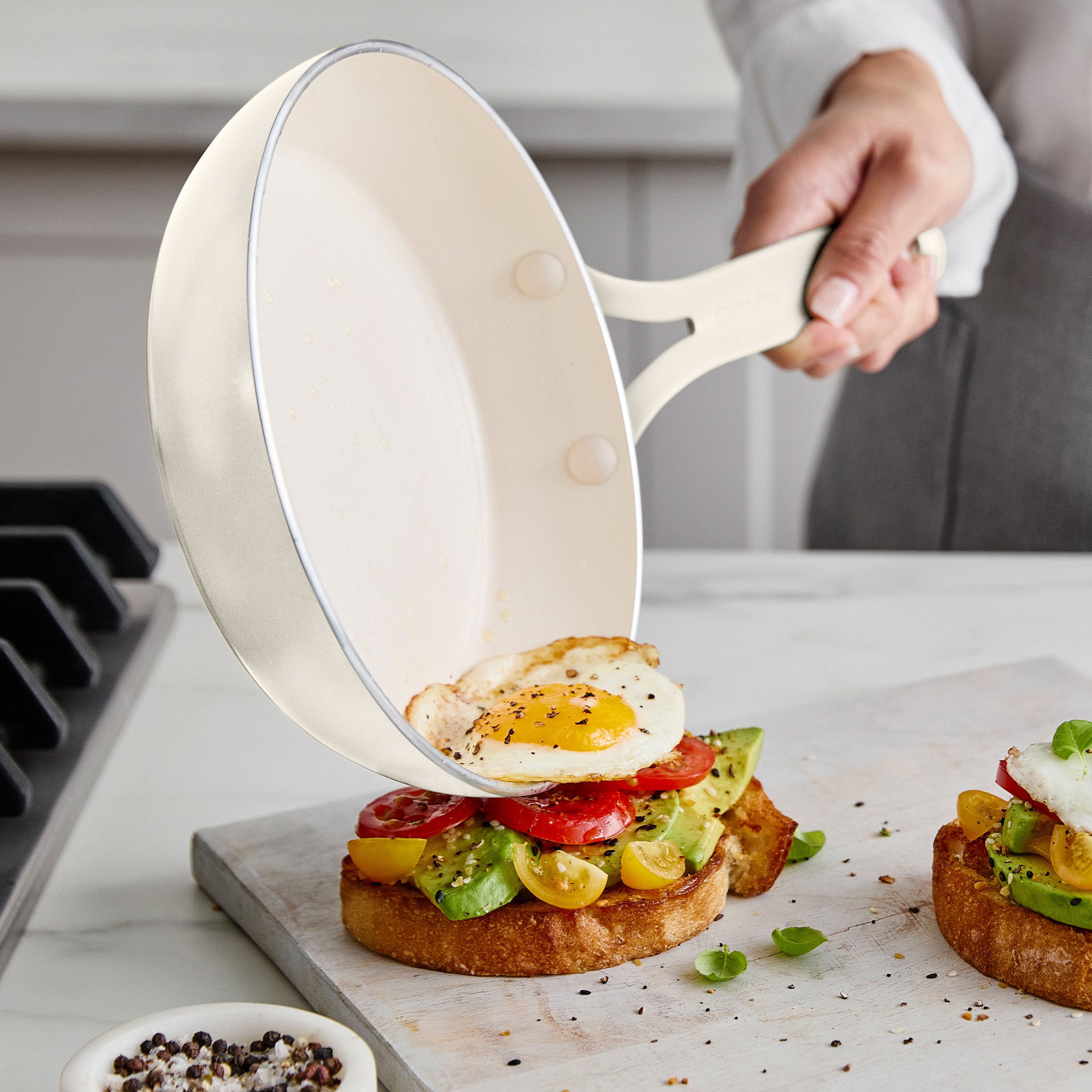 Person using a white frying pan to place an egg on avocado toast on a marble surface.