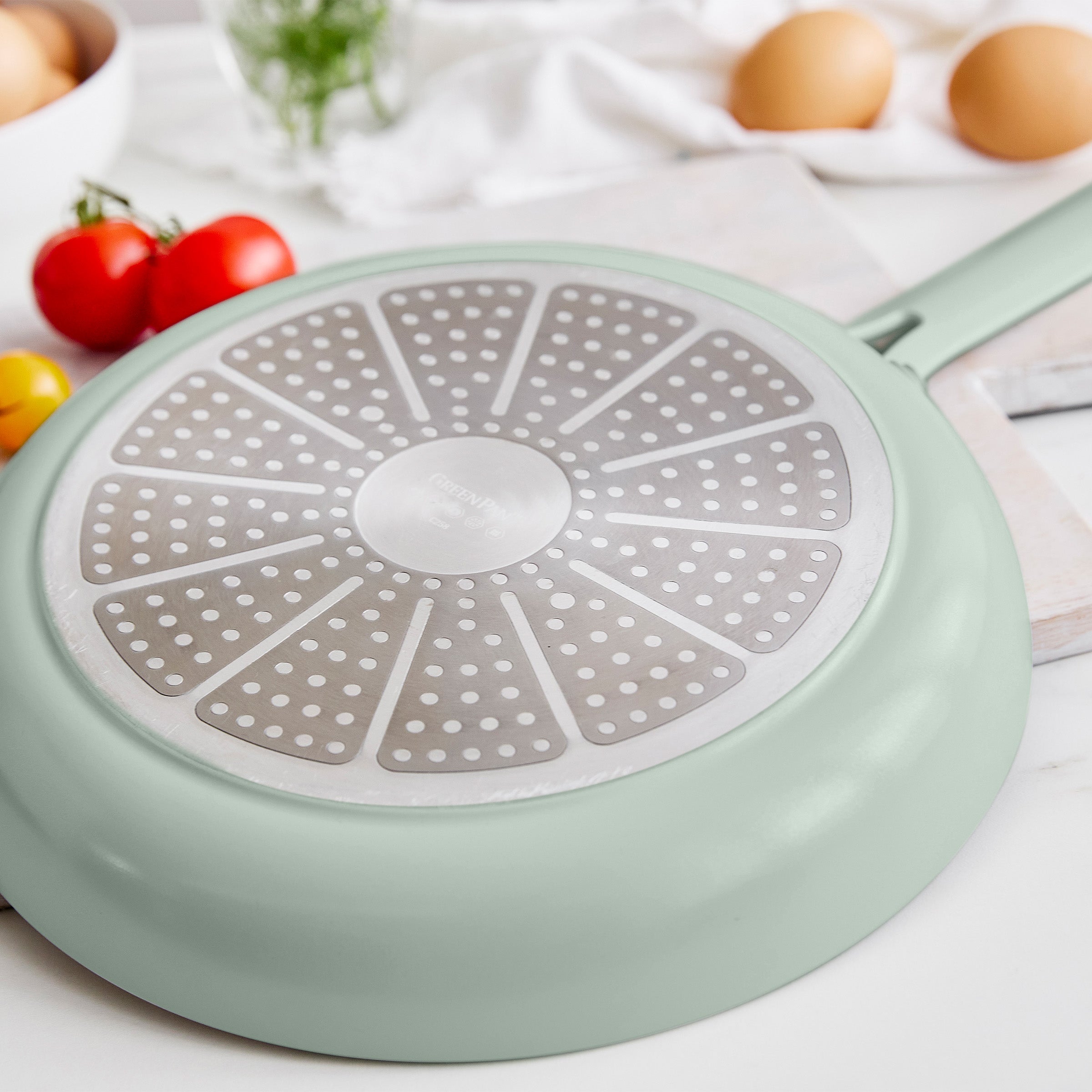 Light green frying pan with a perforated base on a white surface with eggs and tomatoes.